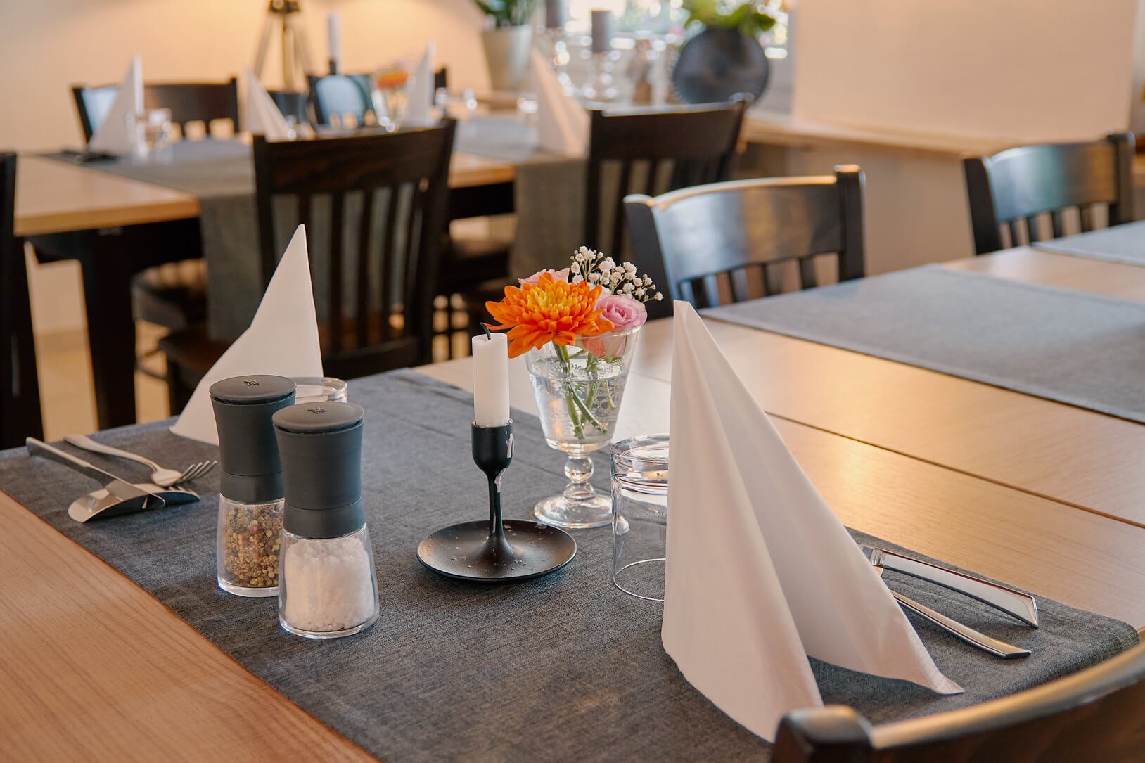 Ein stilvoll gedeckter Esstisch im Restaurant mit weißen Stoffservietten, Kerzen, Gewürzstreuern und einer Blumen Vase.