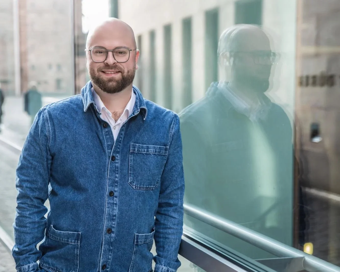 Ein lachender Mann mit Brille, Bart und Glatze, trägt eine blaue Jeansjacke und steht neben einem Fenster auf einem Bürgersteig.
