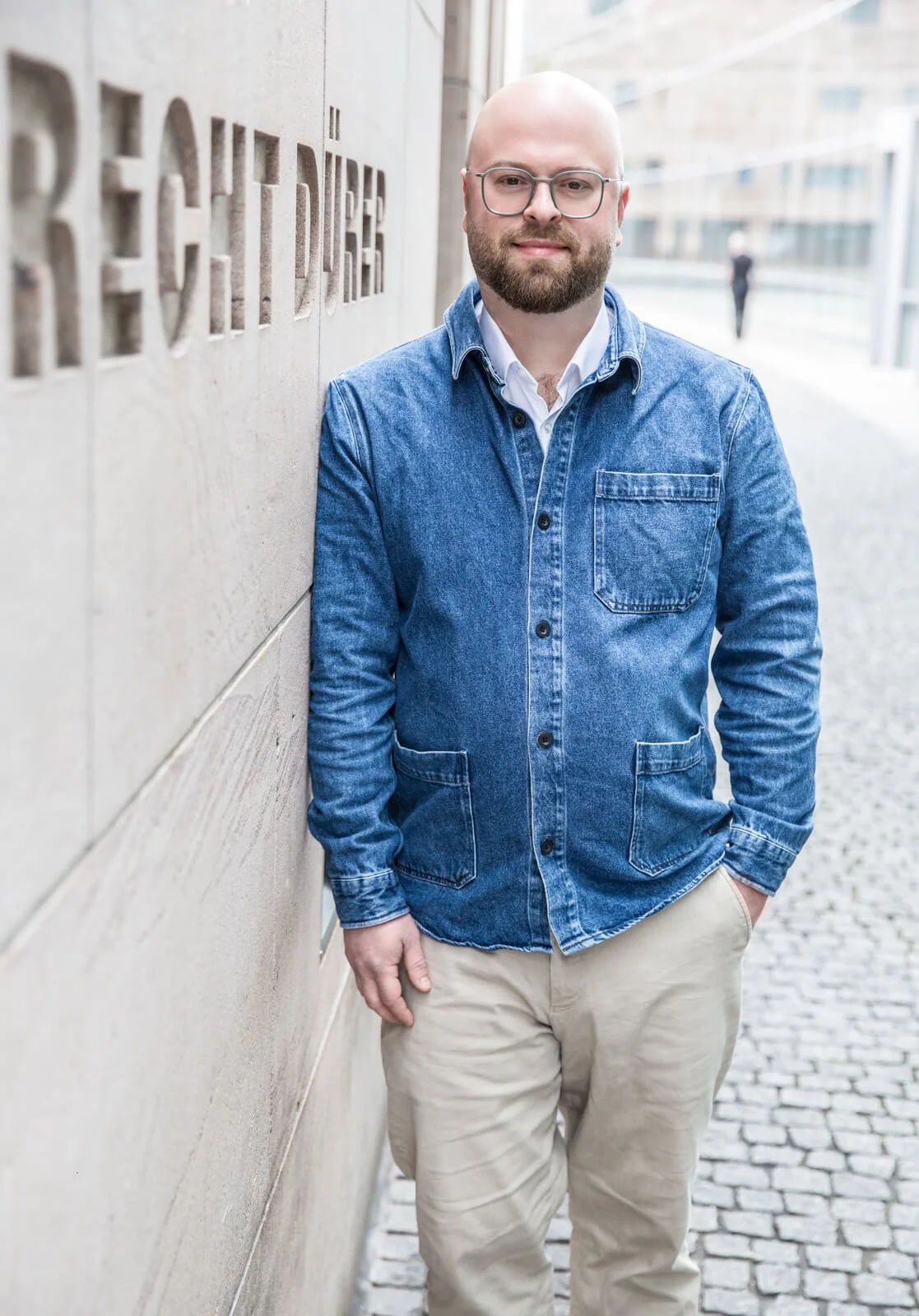 Porträt des Kunsthistorikers Dr. Alexander Racz. Er lehnt an die Mauer des Neuen Museum Nürnberg. Er trägt eine blaue Jeansjacke, ein weisses Hemd und eine beige Hose.