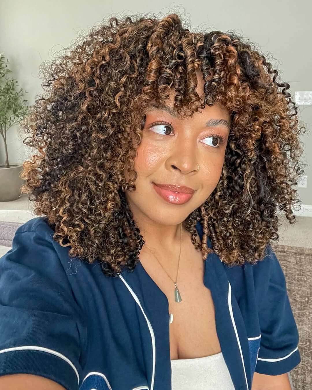A woman with curly, medium-length hair with brown highlights, wearing a navy blue top and a gold necklace, smiling and looking to the side in a living room.
