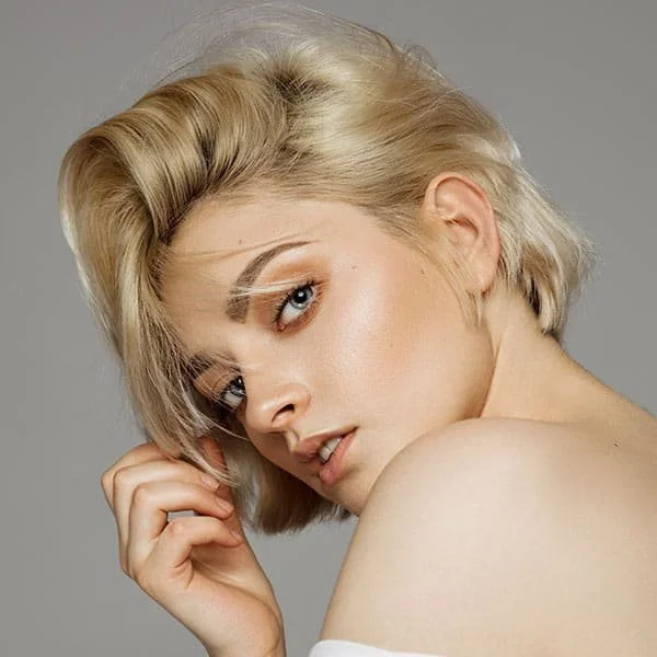 A woman with blonde hair styled in a short side-swept haircut, looking over her shoulder with her hand near her face, against a gray background.