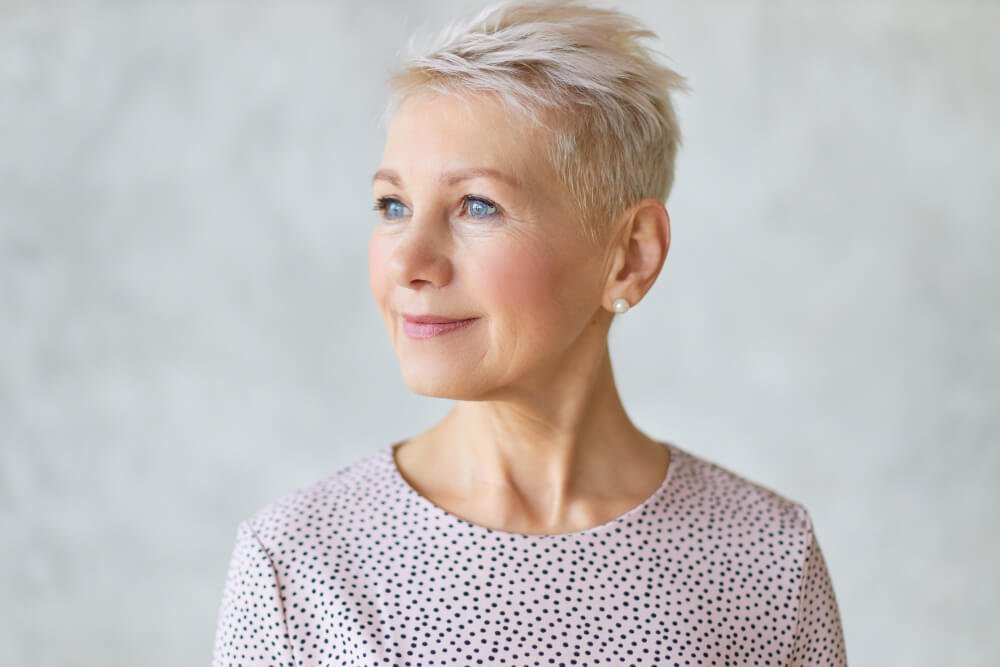 A portrait of an older woman with short, platinum blonde hair and blue eyes, wearing a light, polka-dotted top, looking off to the side against a neutral background.