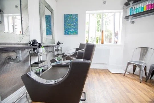 Empty hair salon with two styling chairs facing mirrors, wooden floor, and a shelf with hair color bottles.