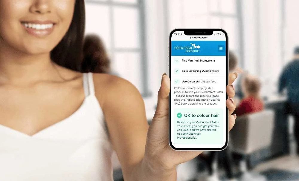 A woman holding a smartphone displaying a hair coloring app in a salon with other people in the background.