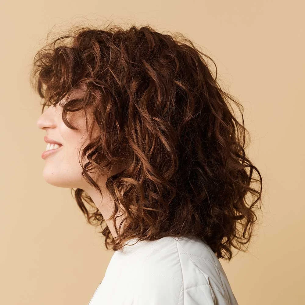 Profile of a woman with shoulder-length, curly, reddish-brown hair, smiling, against a neutral beige background.