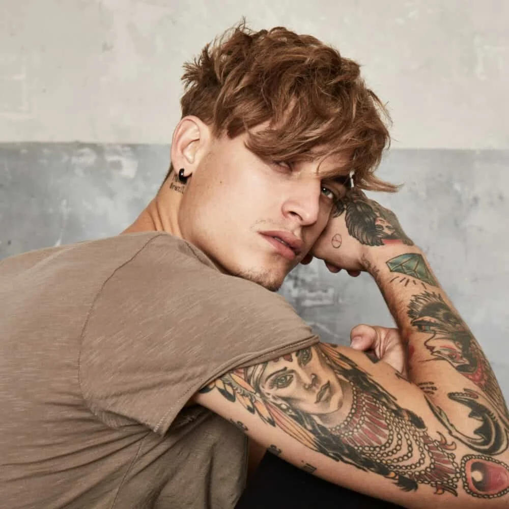 A young man with light skin and brown, wavy hair, wearing a beige T-shirt, is looking over his shoulder at the camera. He has multiple tattoos on his arms and face, including a large portrait of a woman on his forearm, earrings, and a small star tattoo near his eye.