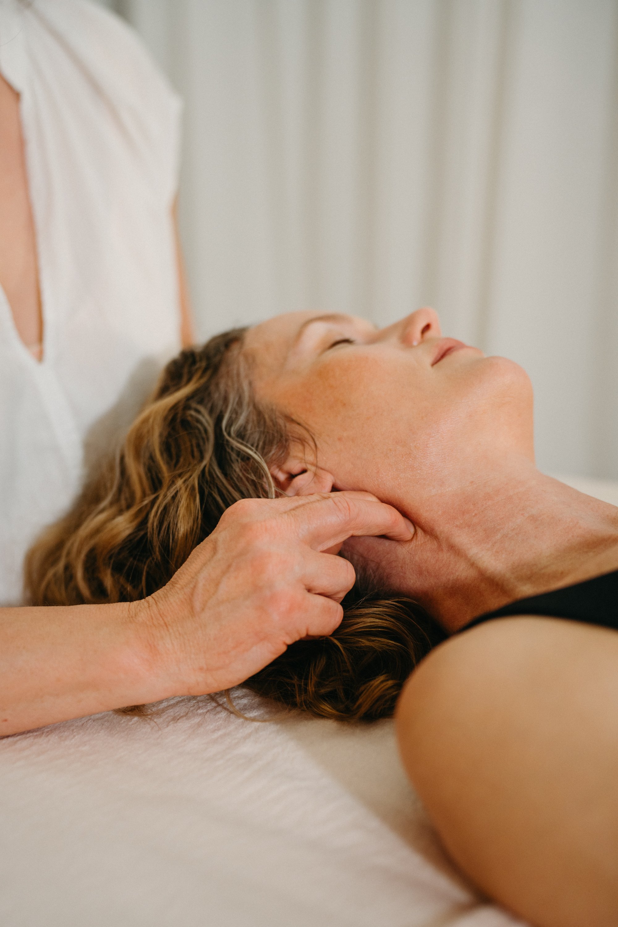 A woman receiving a neck massage while lying down on a massage table, with her eyes closed.