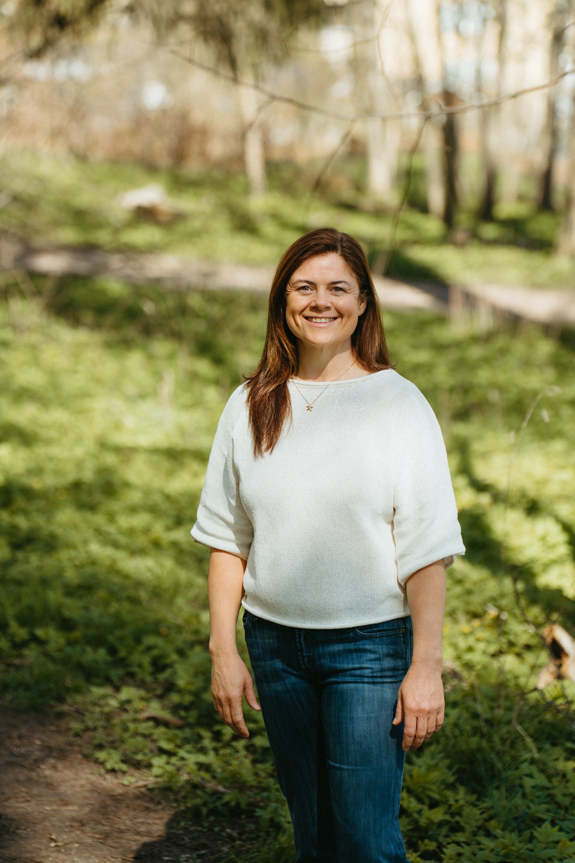 A woman with long brown hair smiling while walking in a wooded park with trees and greenery in the background.