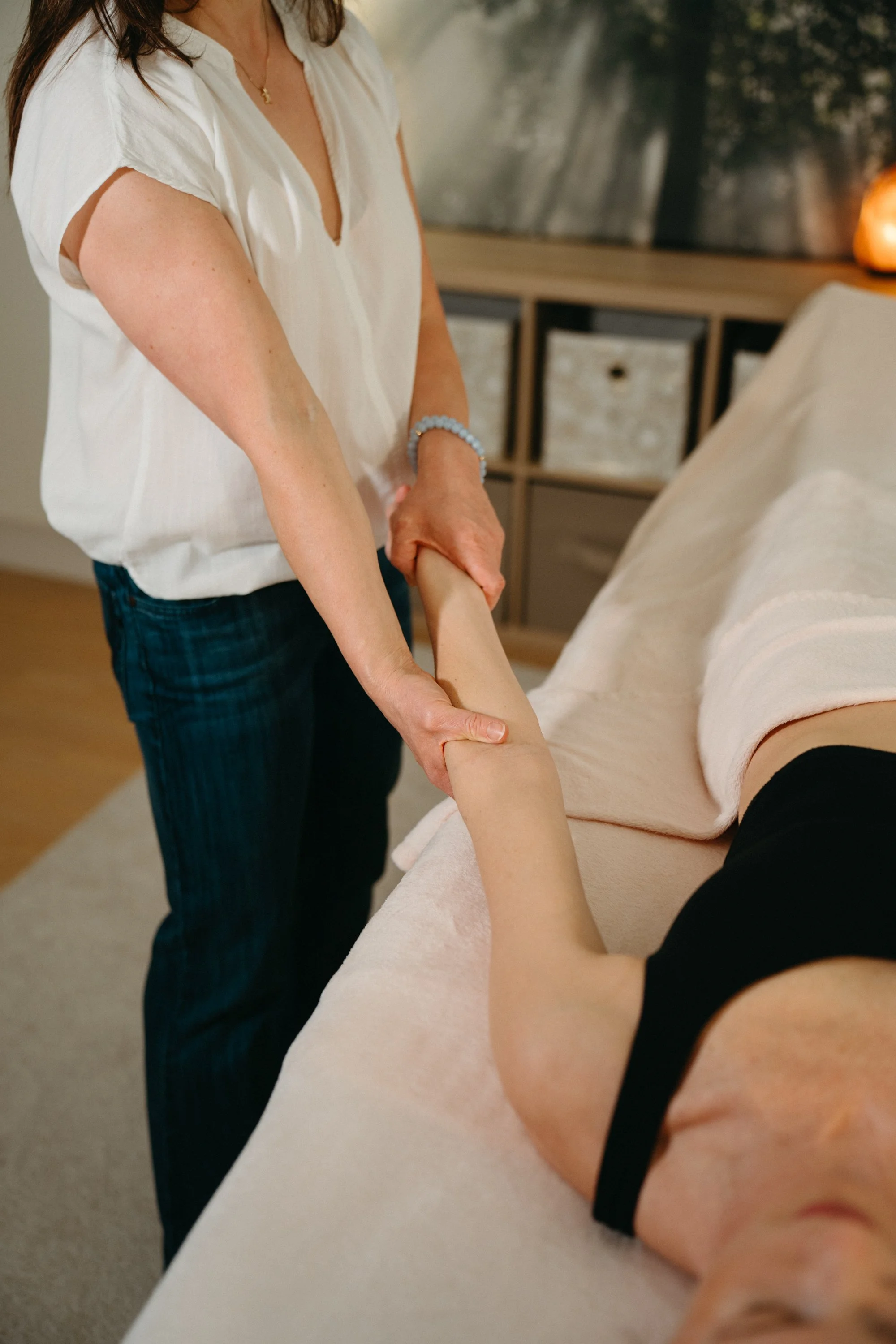 A woman is receiving a massage while lying on a massage table, with a massage therapist gently holding her arm.