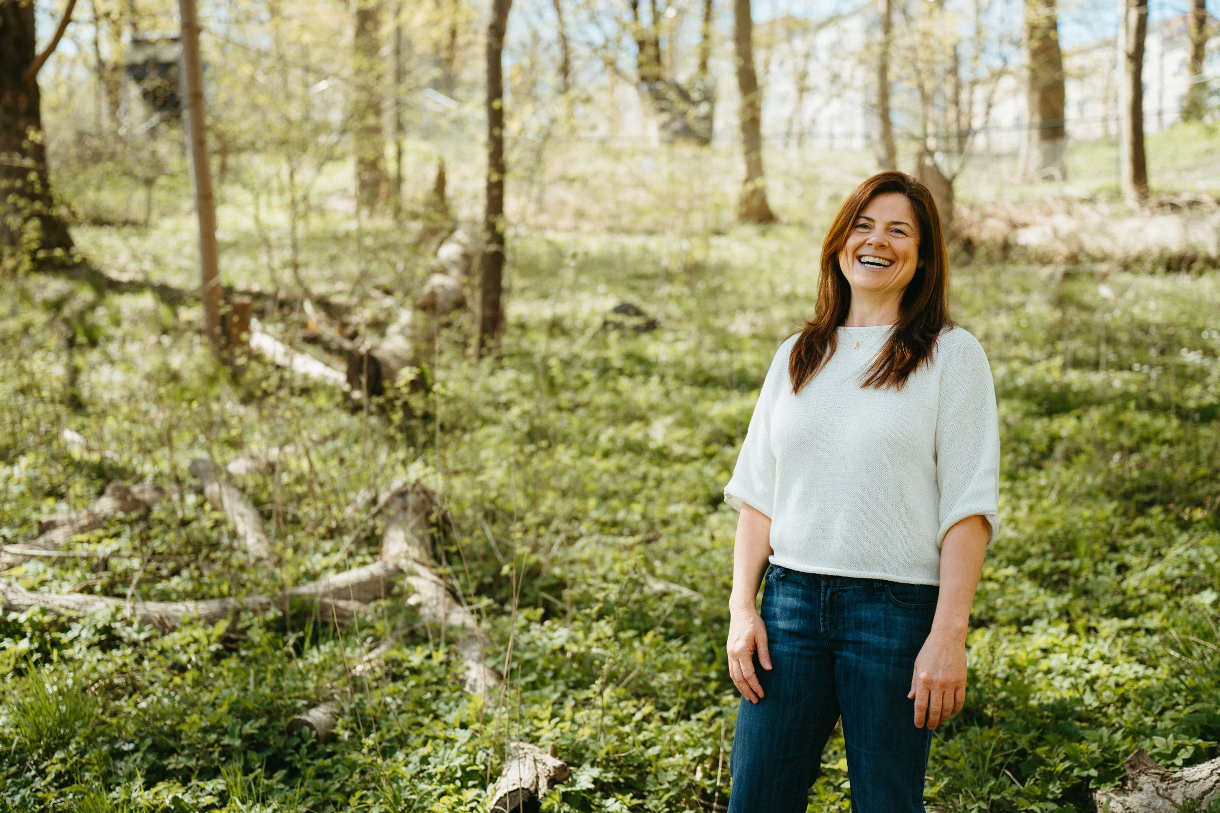 A woman with brown hair smiling in a forest with green trees and foliage.