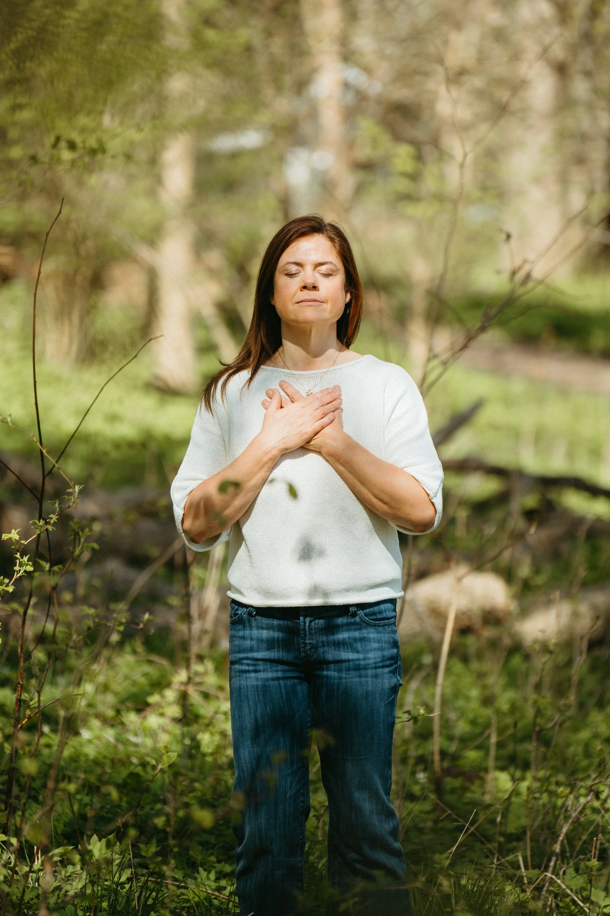 A woman standing outdoors in a wooded area with her eyes closed and hands over her chest, practicing meditation or mindfulness.