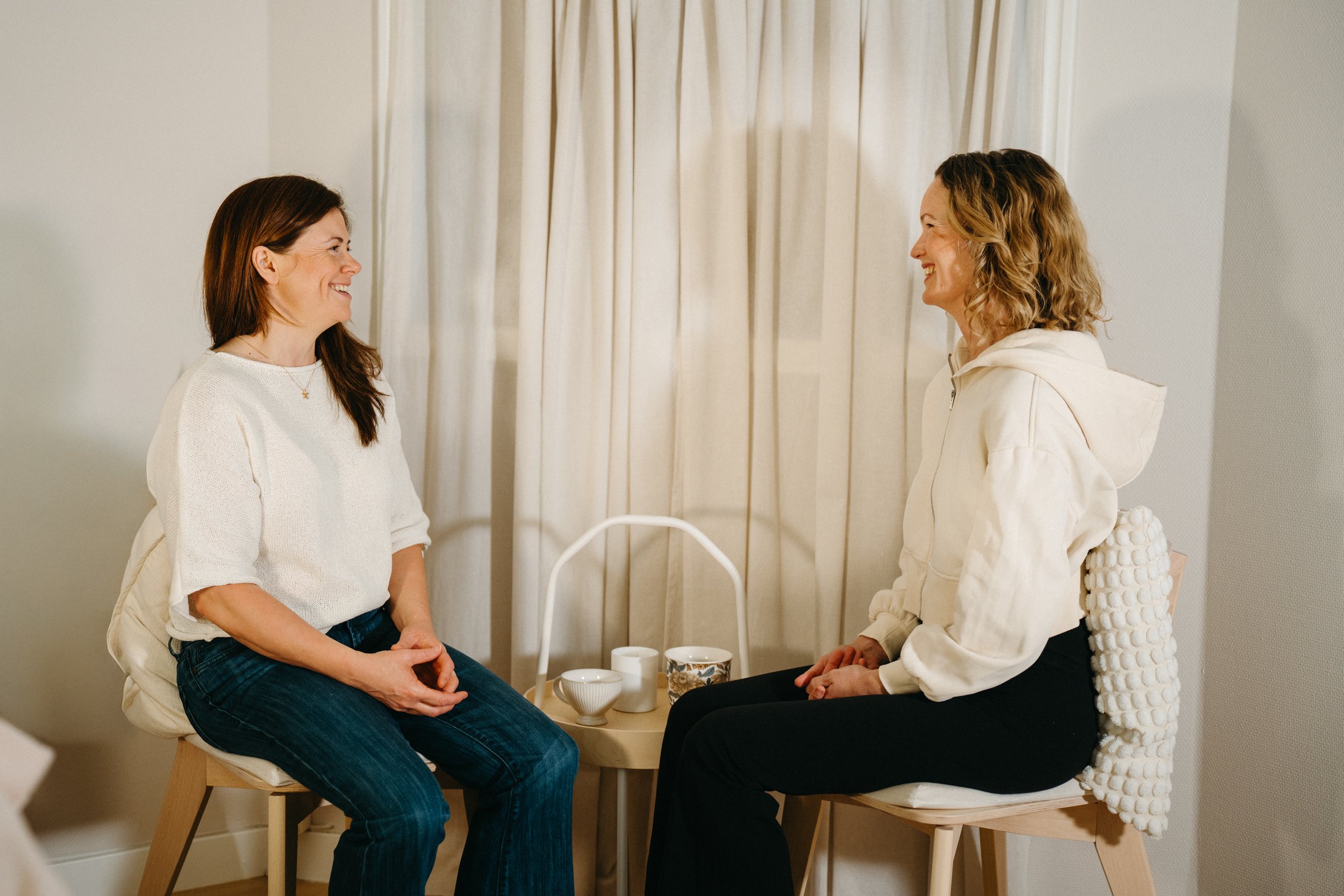 Two women sitting across from each other, smiling and engaging in conversation in a cozy room with cream-colored curtains and tea cups on a small table between them.