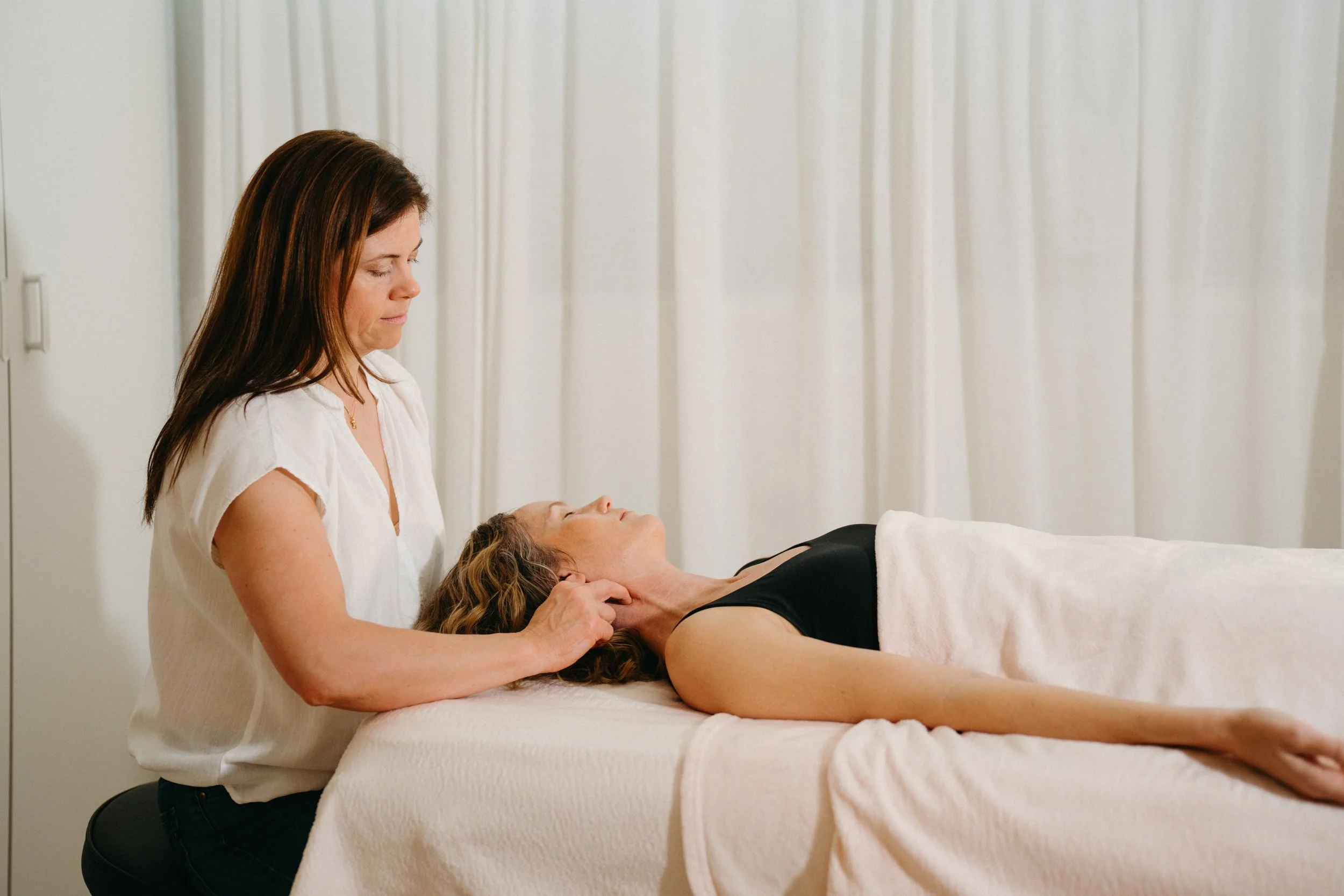 A woman lying on her back on a massage table with a towel draped over her body, receiving a neck and shoulder massage from a massage therapist in a clinical setting with curtains in the background.