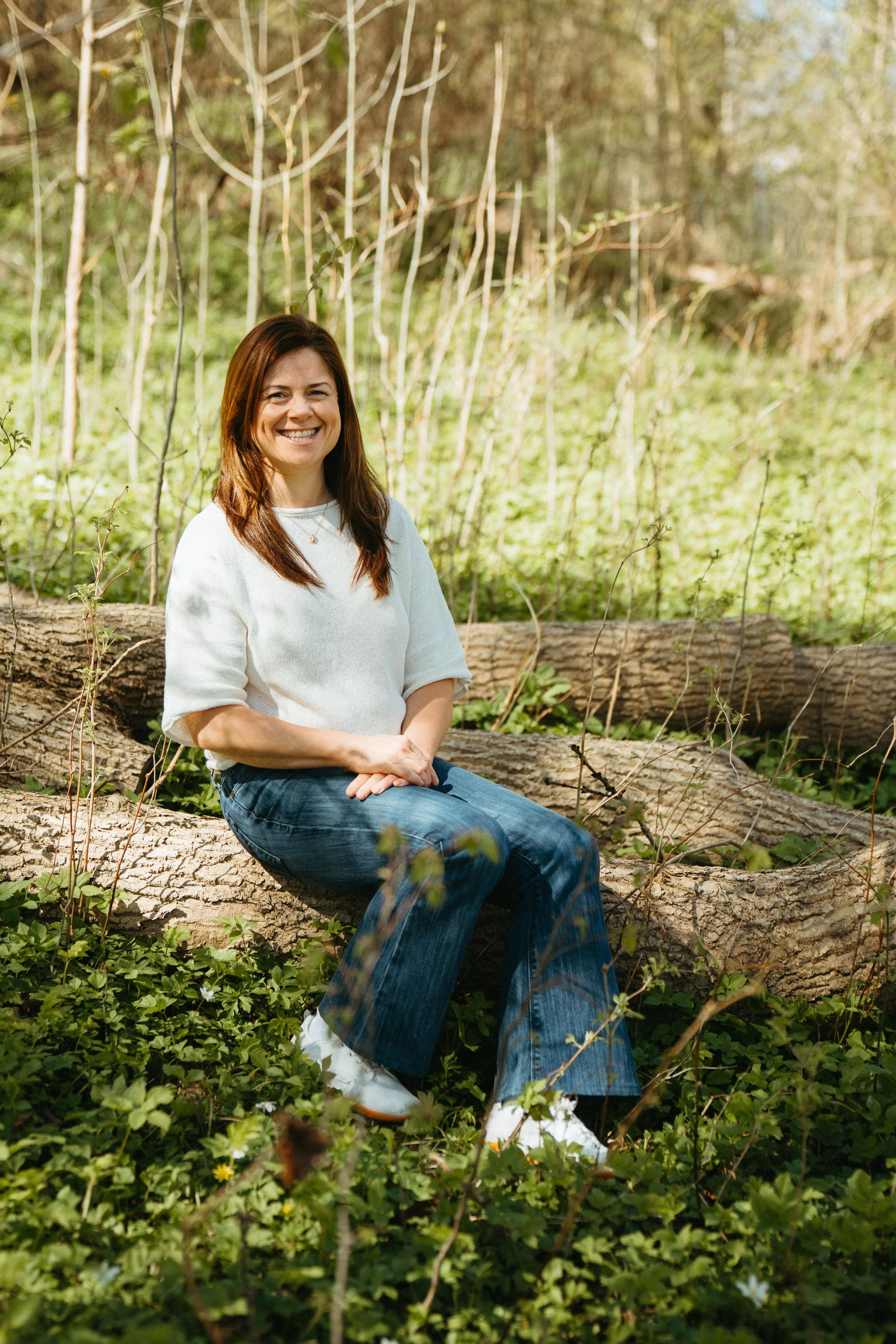 A woman sitting on a fallen tree trunk in a forest, smiling at the camera, surrounded by green plants and small flowers.