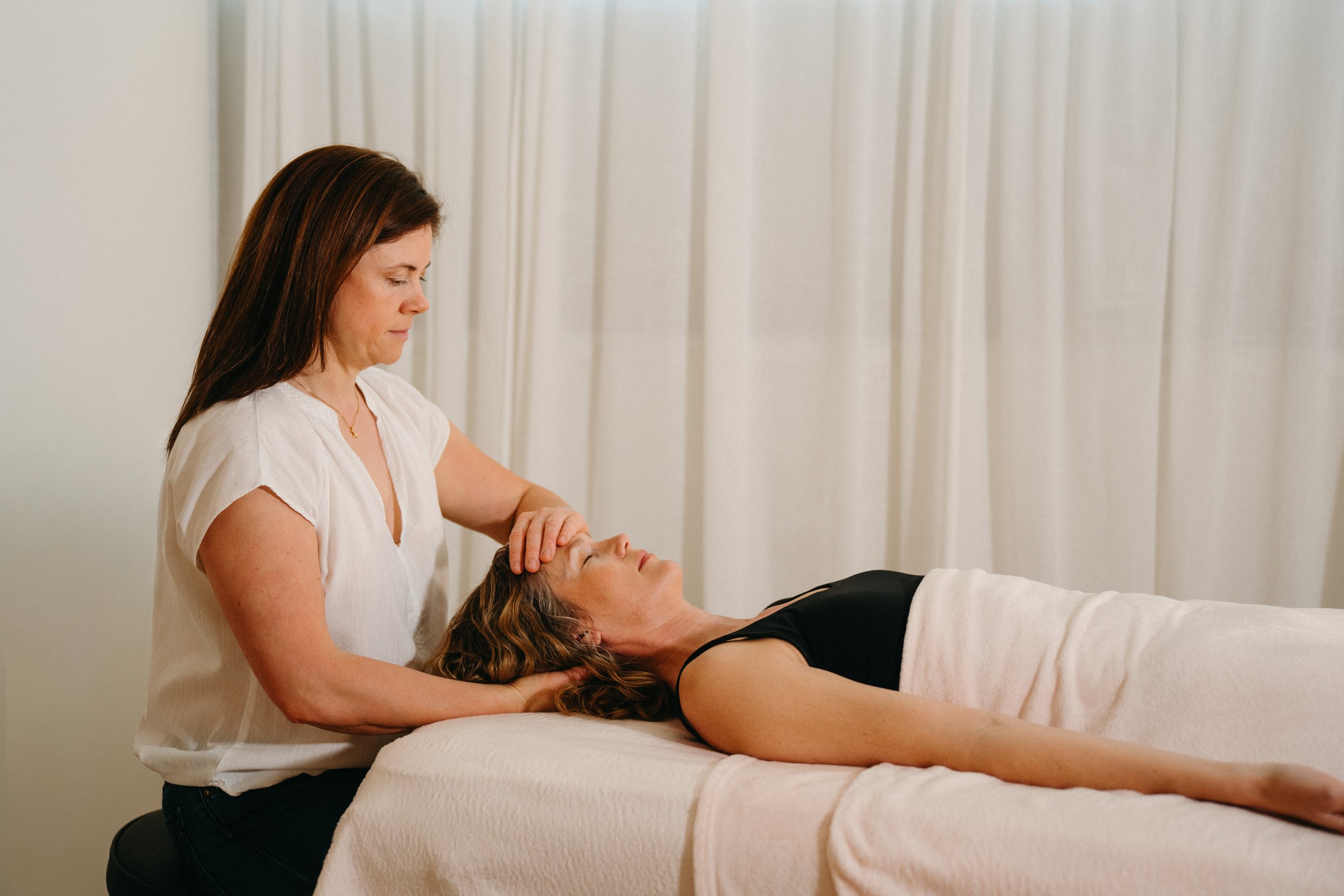 A woman lying on a massage table with her eyes closed, receiving a head massage from another woman in a white shirt, in a room with white curtains.