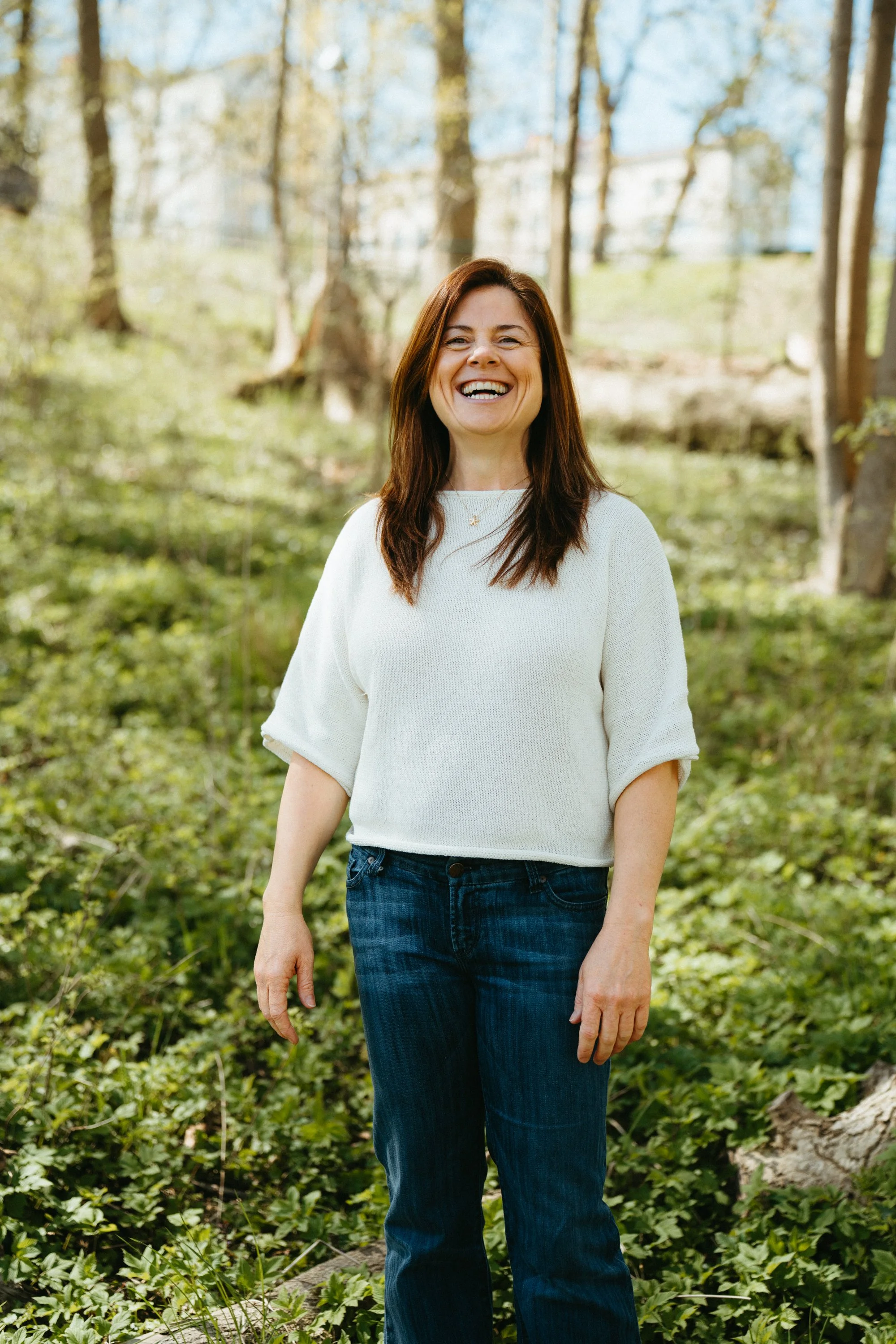 A woman with shoulder-length brown hair smiling outdoors in a park with trees and green foliage in the background.
