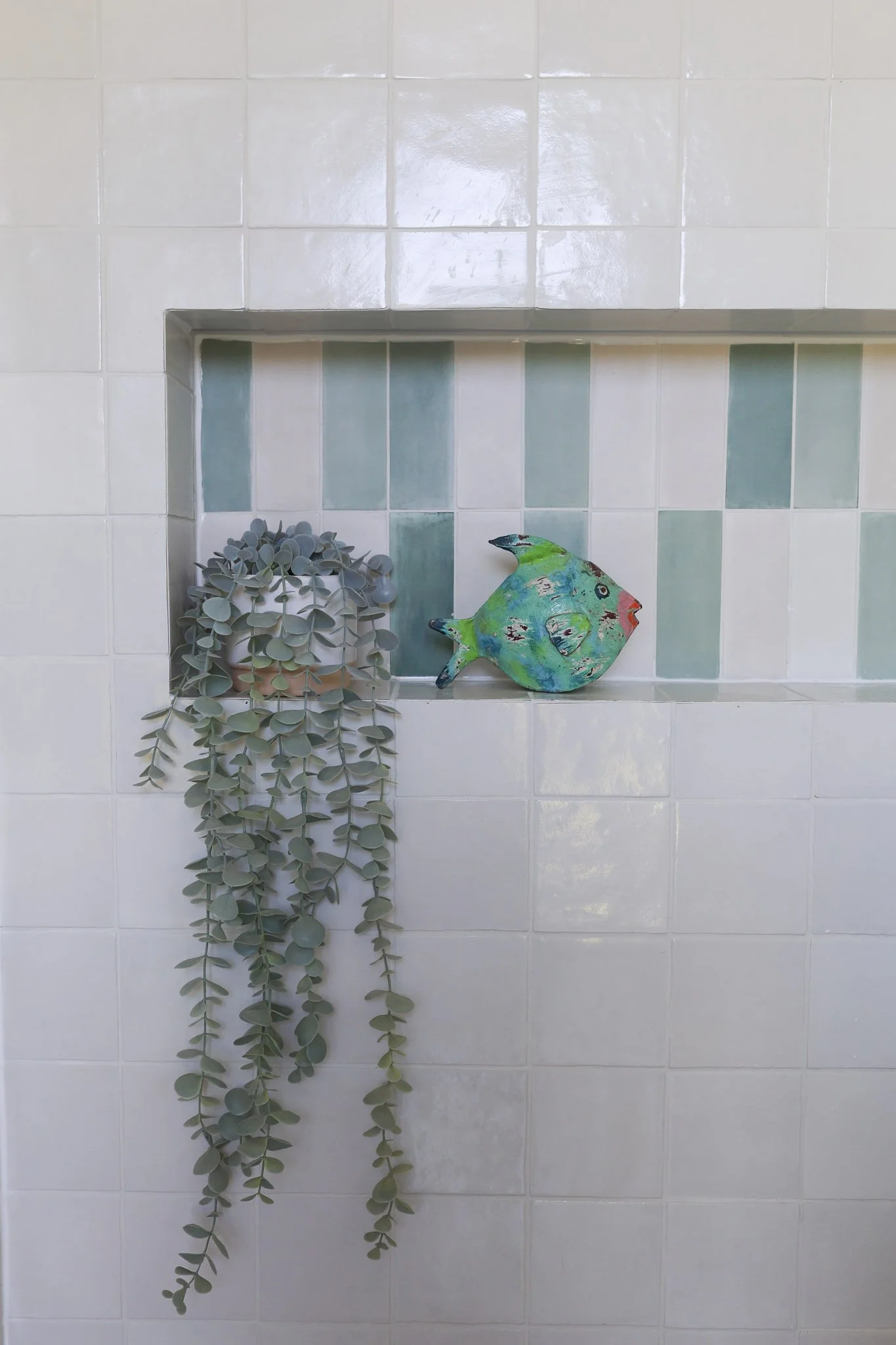 A small artificial green and blue fish decoration with black and red markings on a shelf, next to a potted plant with cascading round gray-green leaves, against a background of white tiles with a light green tile accent.