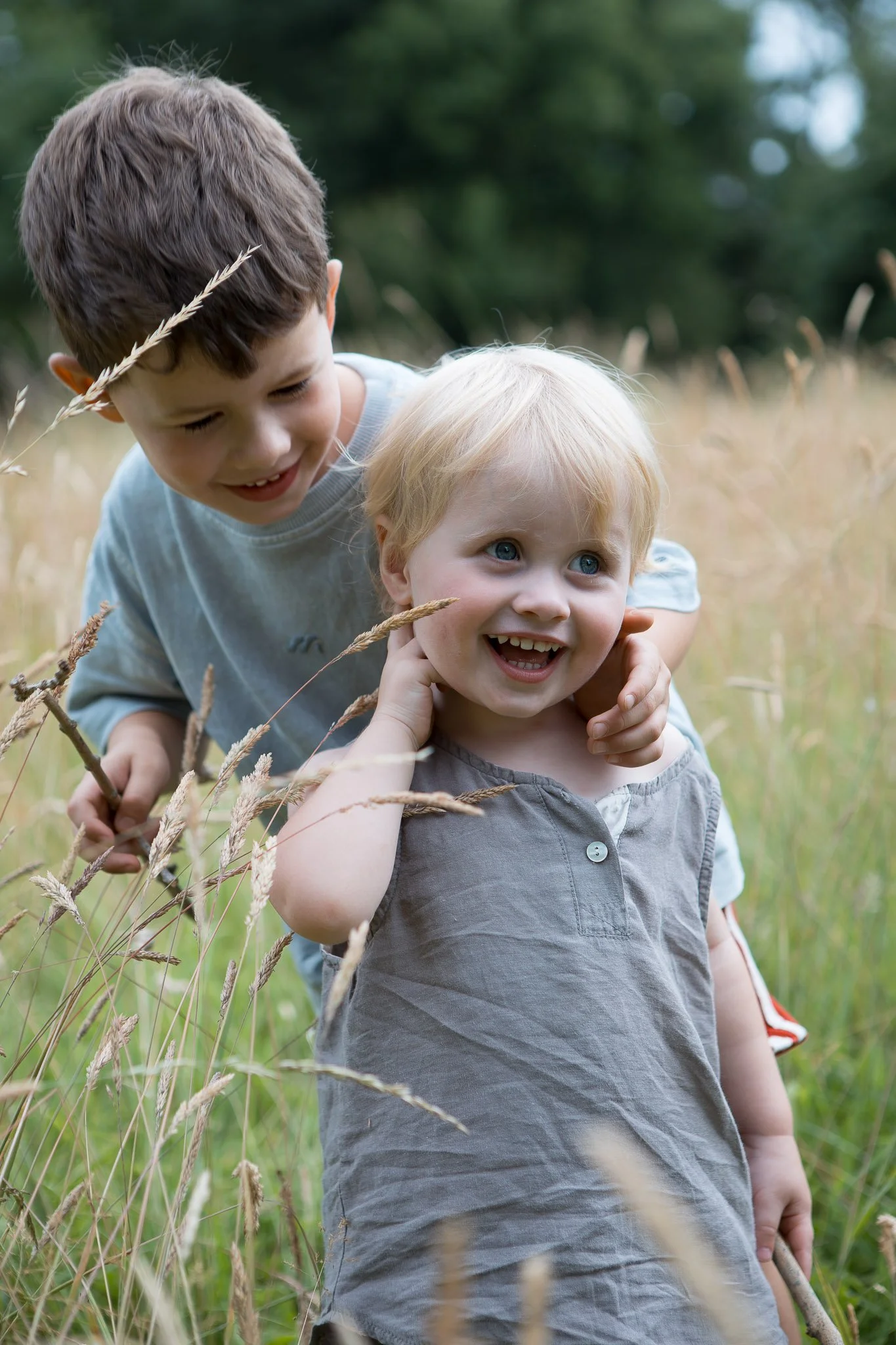 Two young boys playing and smiling in a grassy field, surrounded by tall grasses and trees in the background.