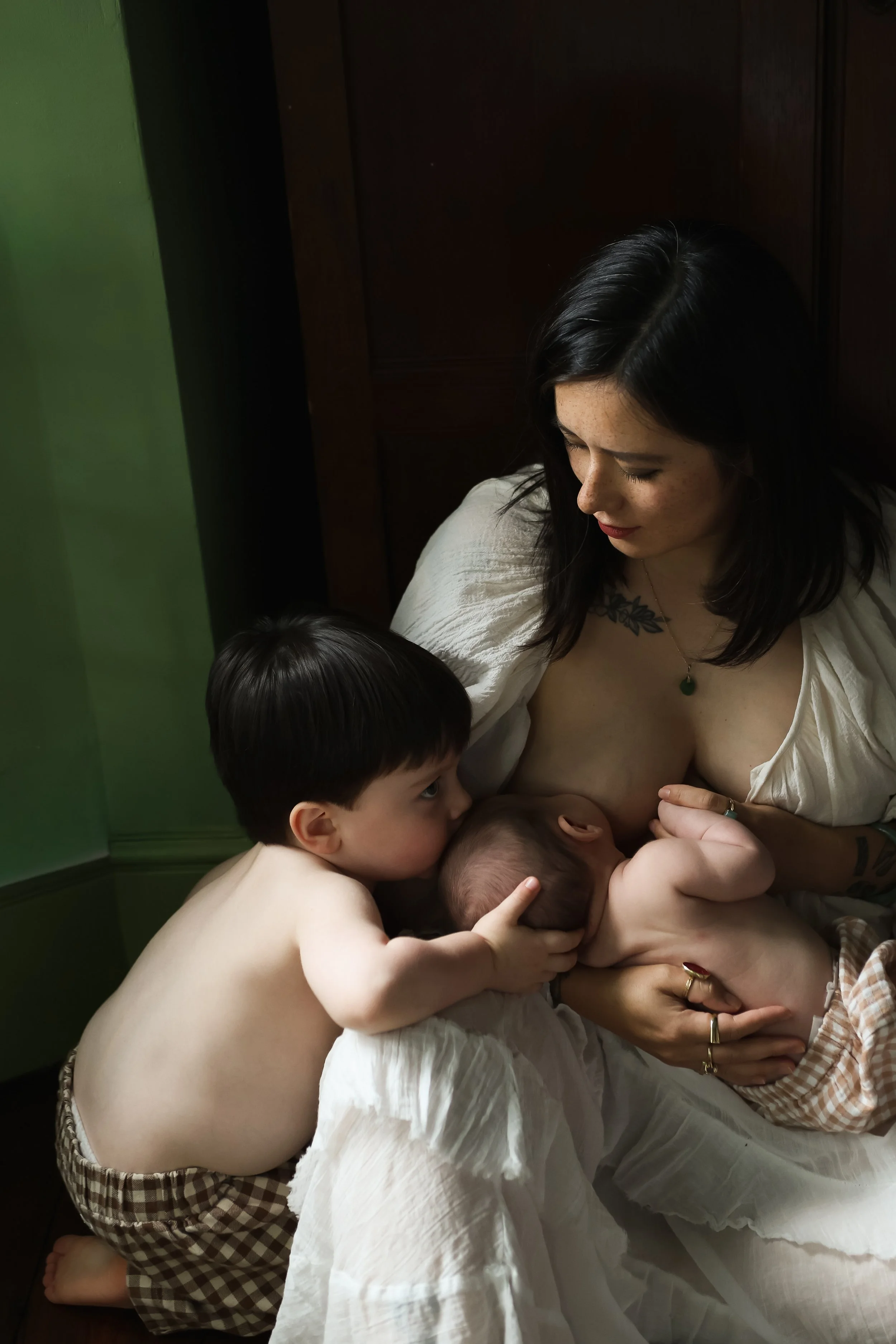 A woman with black hair and a tattoo on her chest feeds four young children a baby bottle in an indoor setting with green wall and wooden furniture.