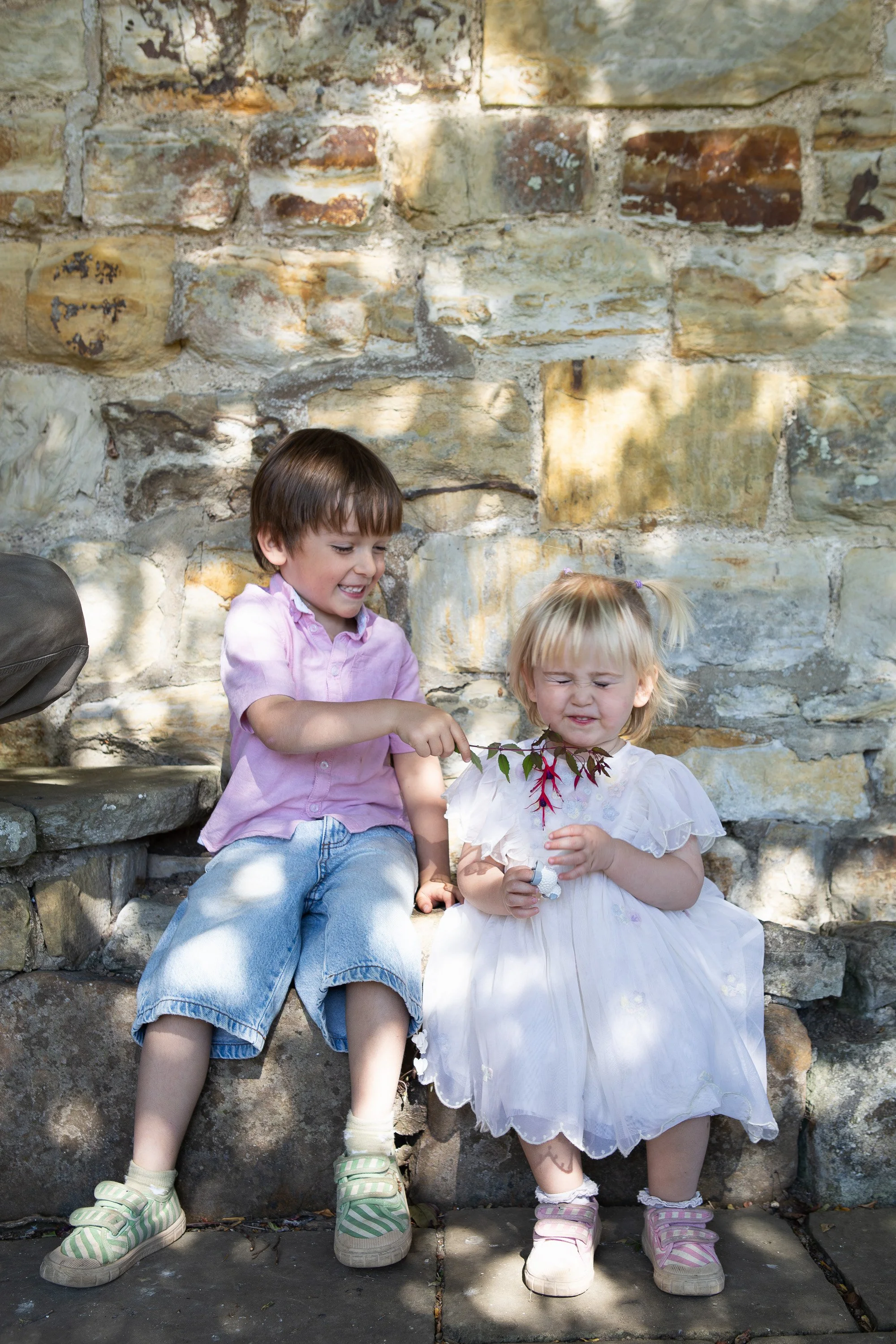 Two young girls sitting on stone steps against a stone wall, holding a branch with leaves. The girl on the left is smiling and wearing a pink shirt and striped sandals. The girl on the right has a pained expression and is wearing a white dress with pink sandals.