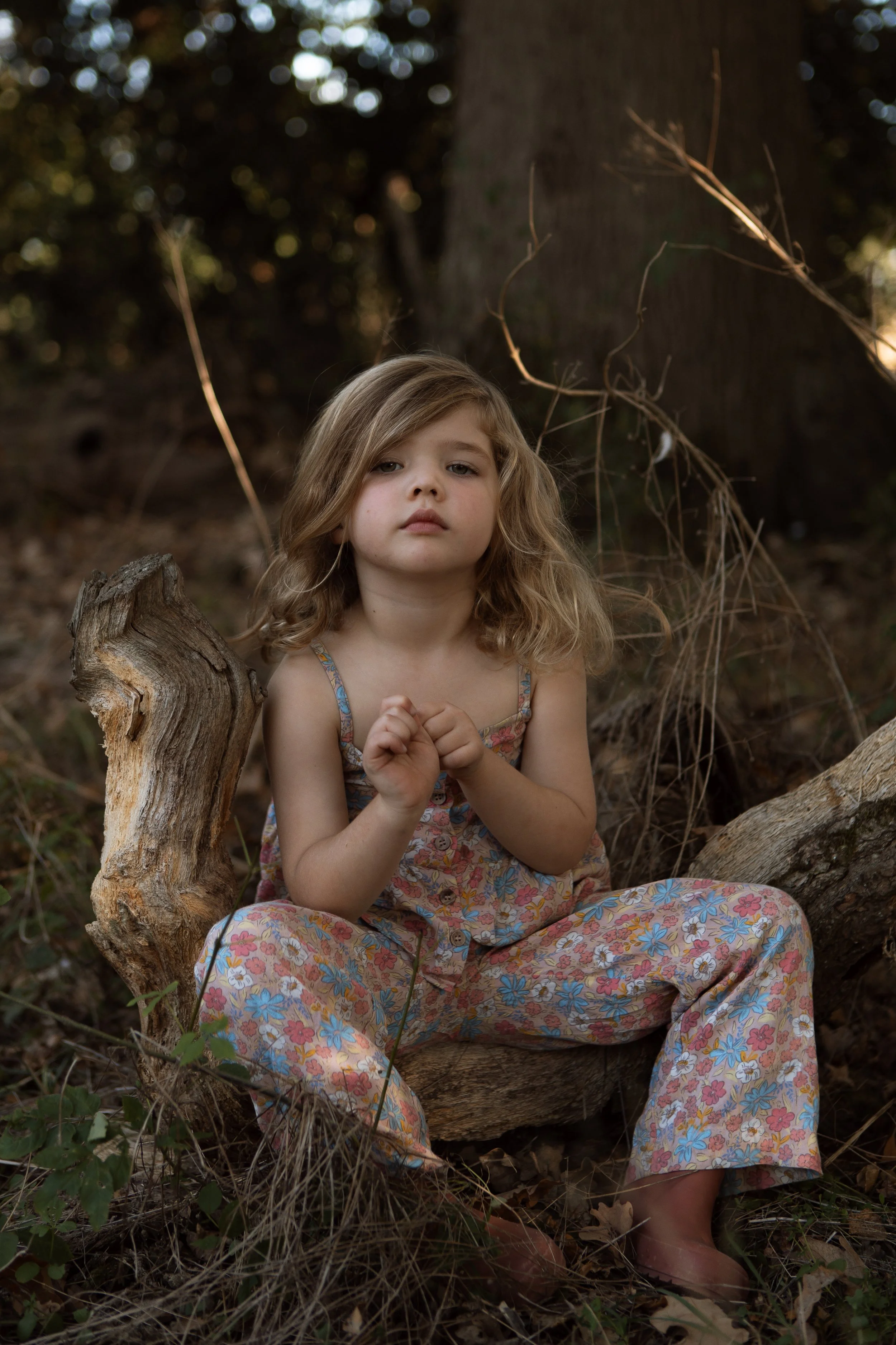 A young girl with blonde wavy hair sitting outdoors near a large tree trunk, surrounded by dried grass and leaves, with a dark forest background.