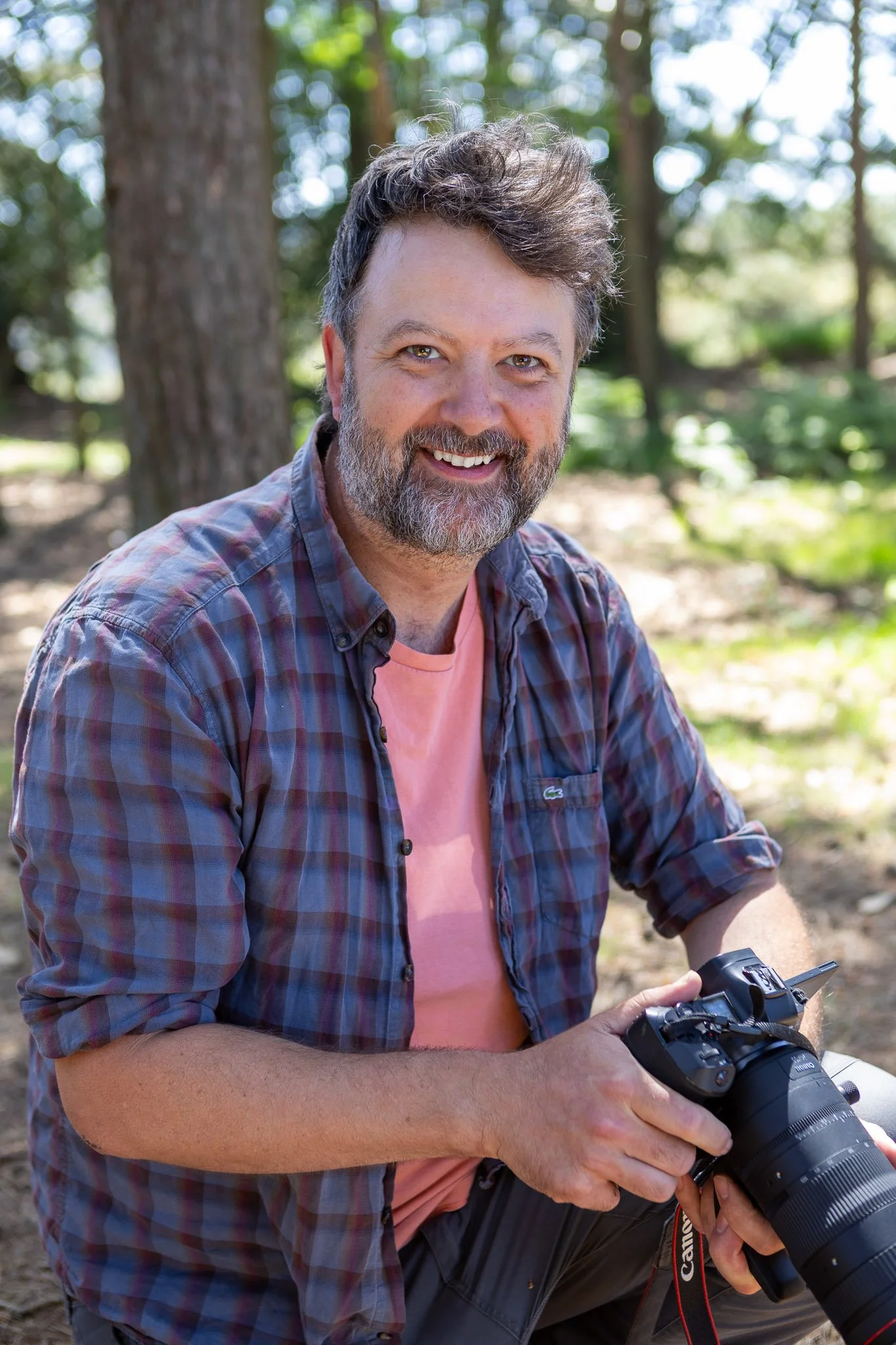 A smiling man with a beard and mustache wearing a plaid shirt sitting outdoors with a camera in his hands, surrounded by trees and greenery.