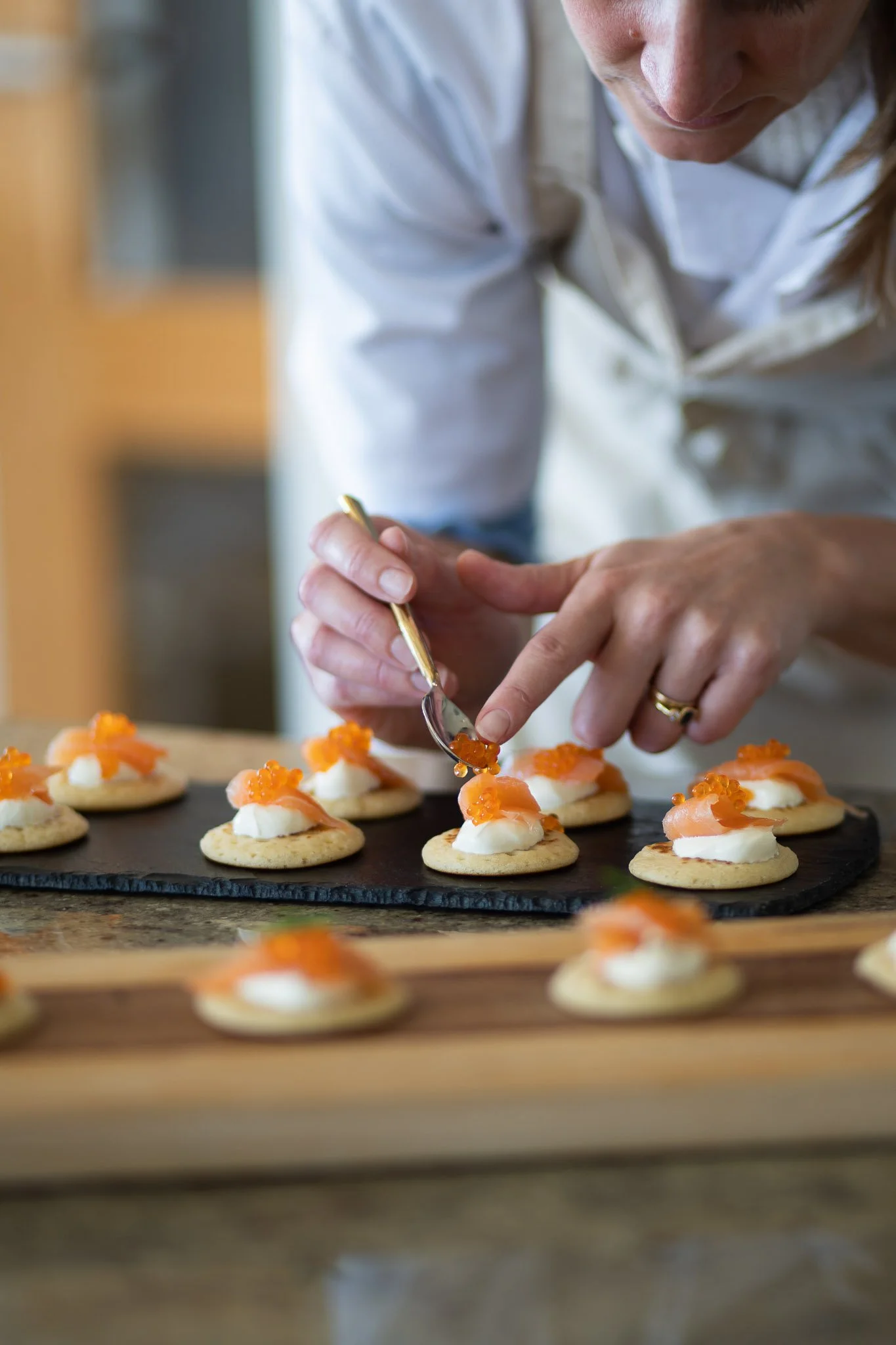 A chef in a white coat is decorating small round appetizers with orange fish roe on top of a dollop of white sauce, arranged on a black slate serving platter.