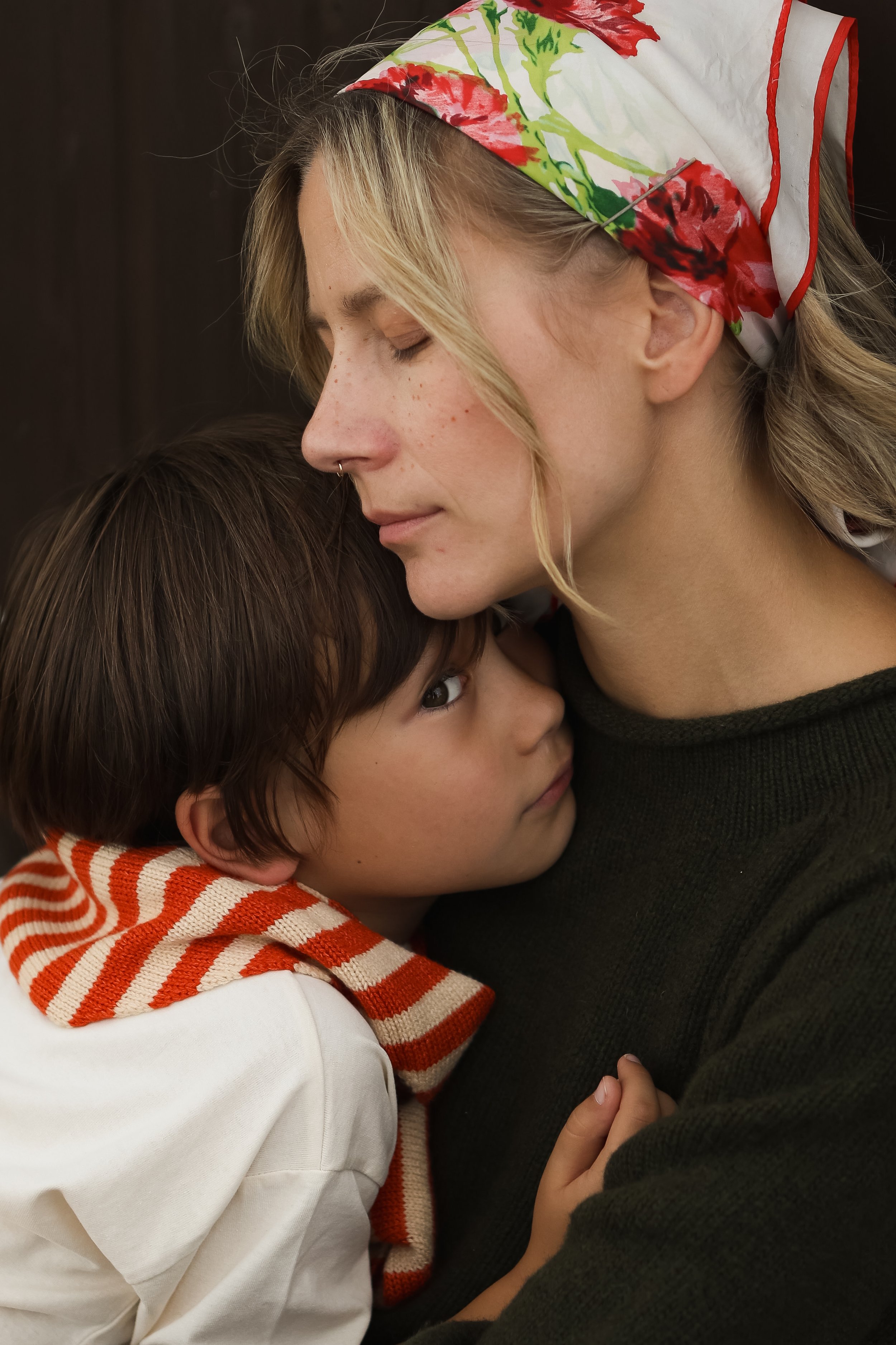 A woman with blonde hair wearing a colorful headscarf gently resting her forehead against a young boy with dark brown hair and a striped red and white hoodie, in a close, intimate embrace.