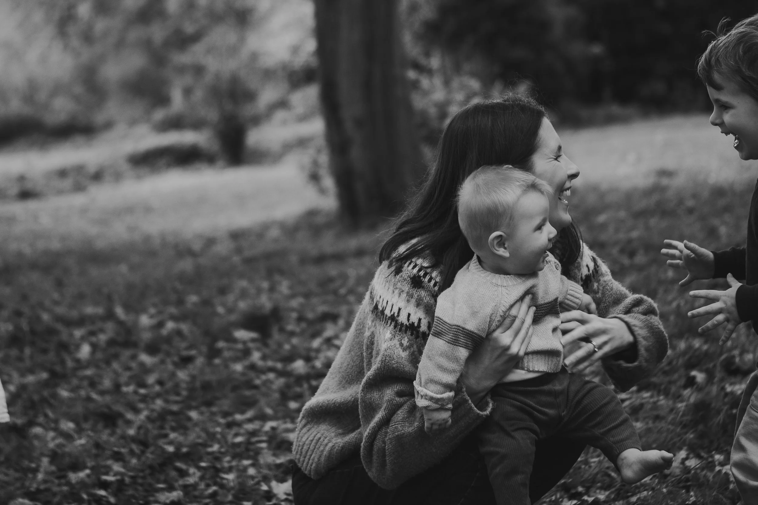 A black-and-white photo of a family of four, a mother and three children, smiling and sharing a moment outdoors in a park or wooded area, with trees and foliage in the background.