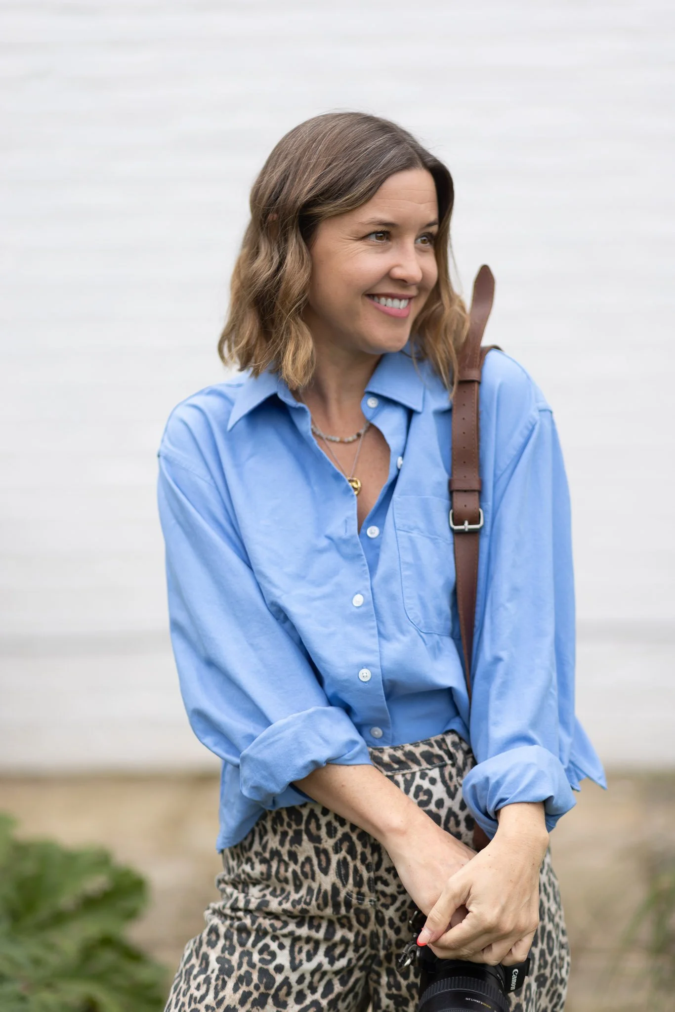 A woman with brown hair, wearing a blue button-up shirt and leopard-print pants, holding a camera at her waist, smiling outdoors against a white background.