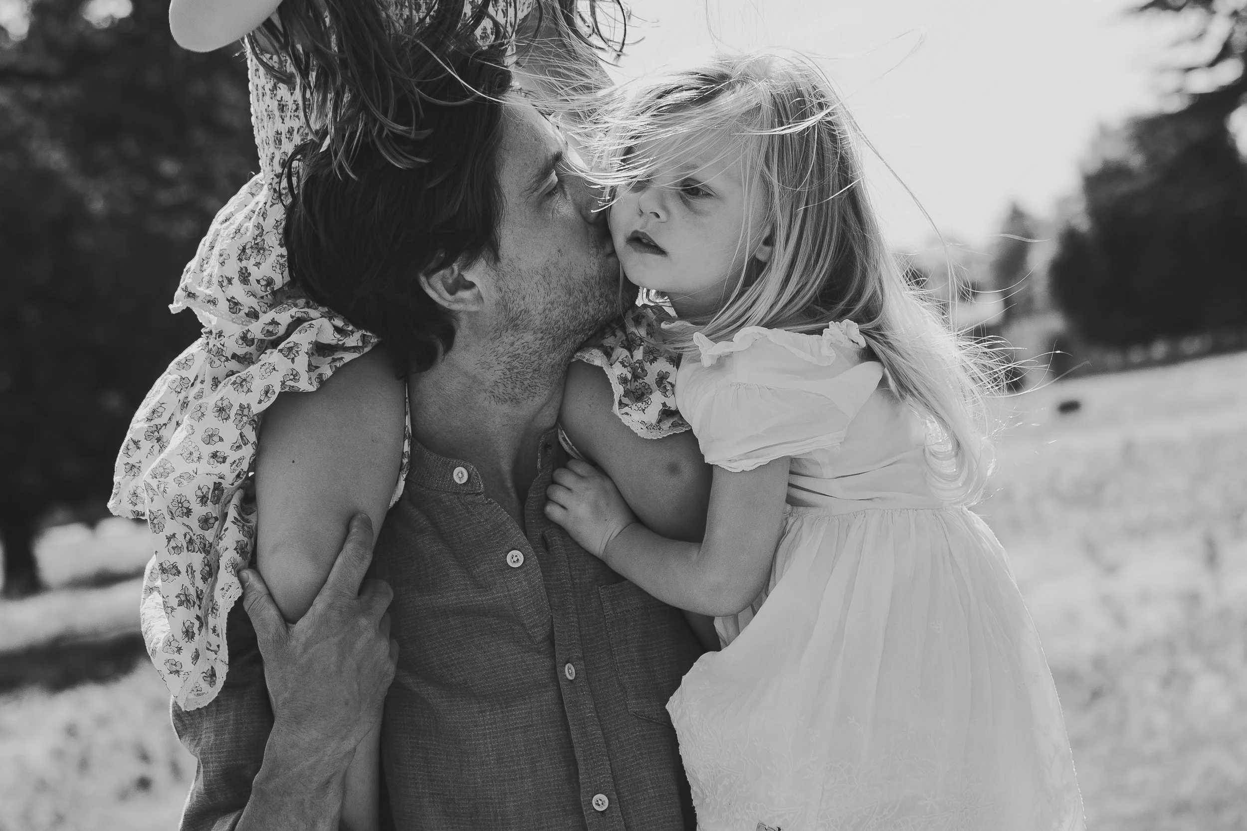 A man holding a young girl in his arms outdoors, with a blurred background of trees and sky, black and white photograph.