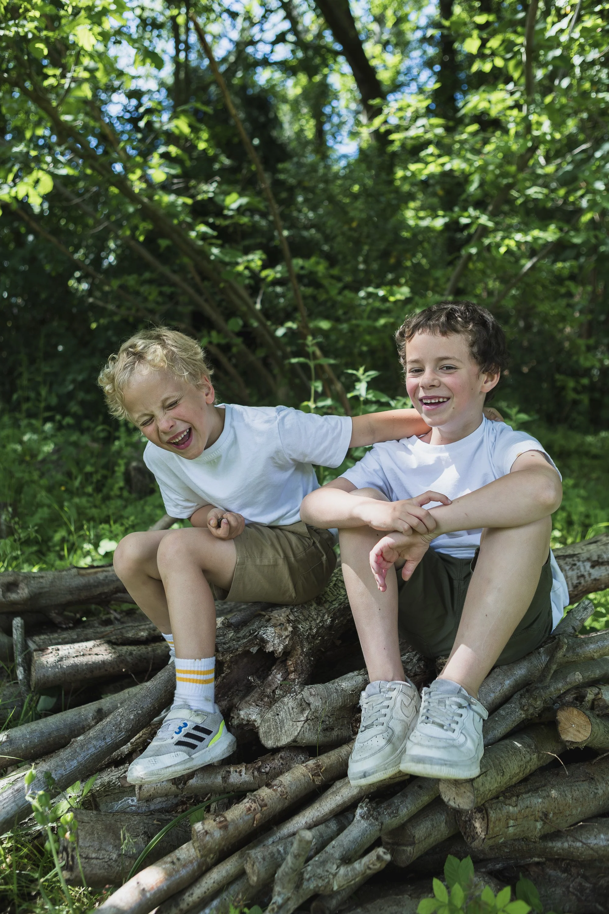 Two young boys sitting on a log in a wooded outdoor area, smiling and playing together, wearing white T-shirts and shorts.
