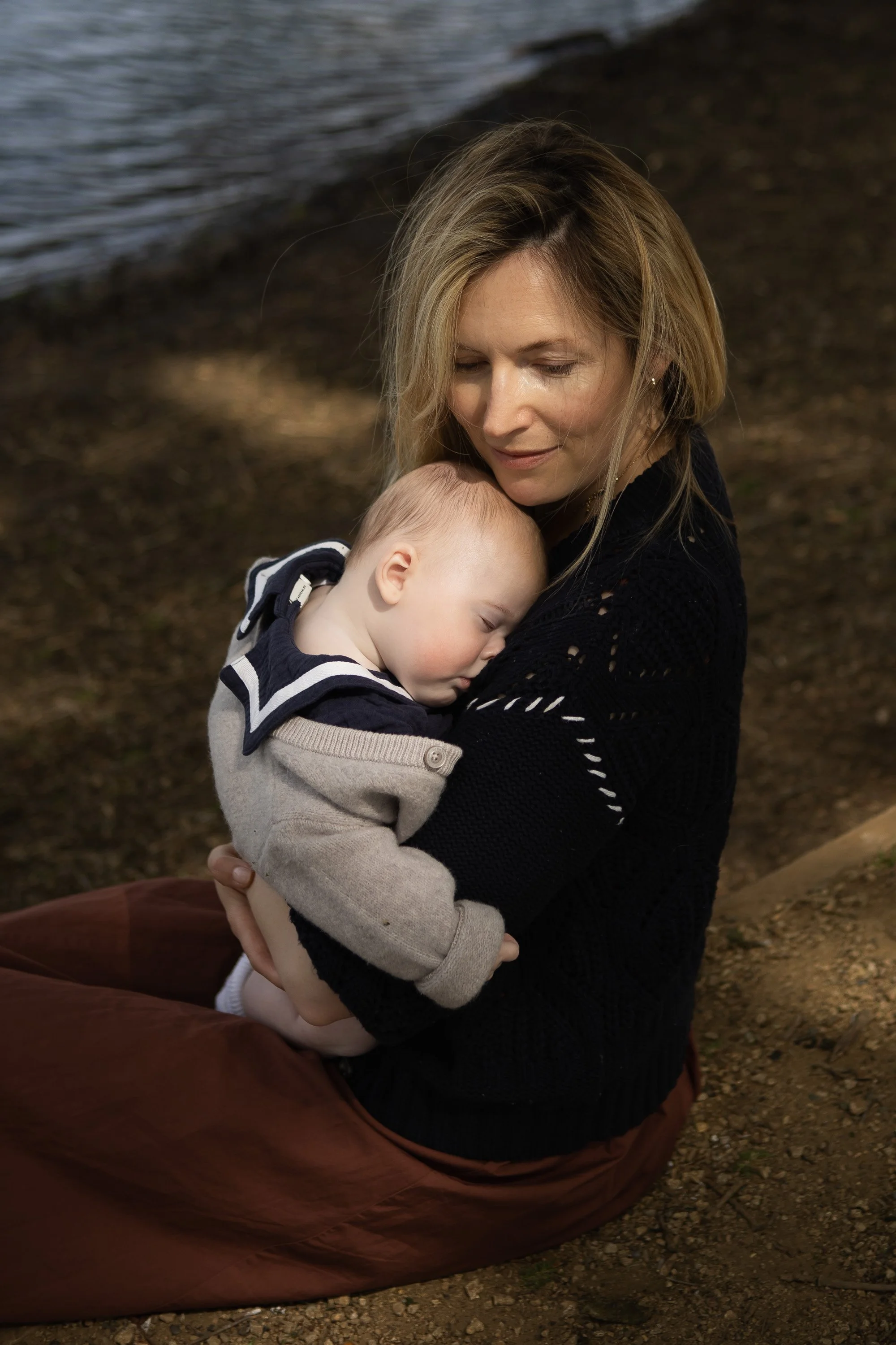 A woman with blonde hair holding a sleeping young boy with light hair and closed eyes, outdoors near a body of water.