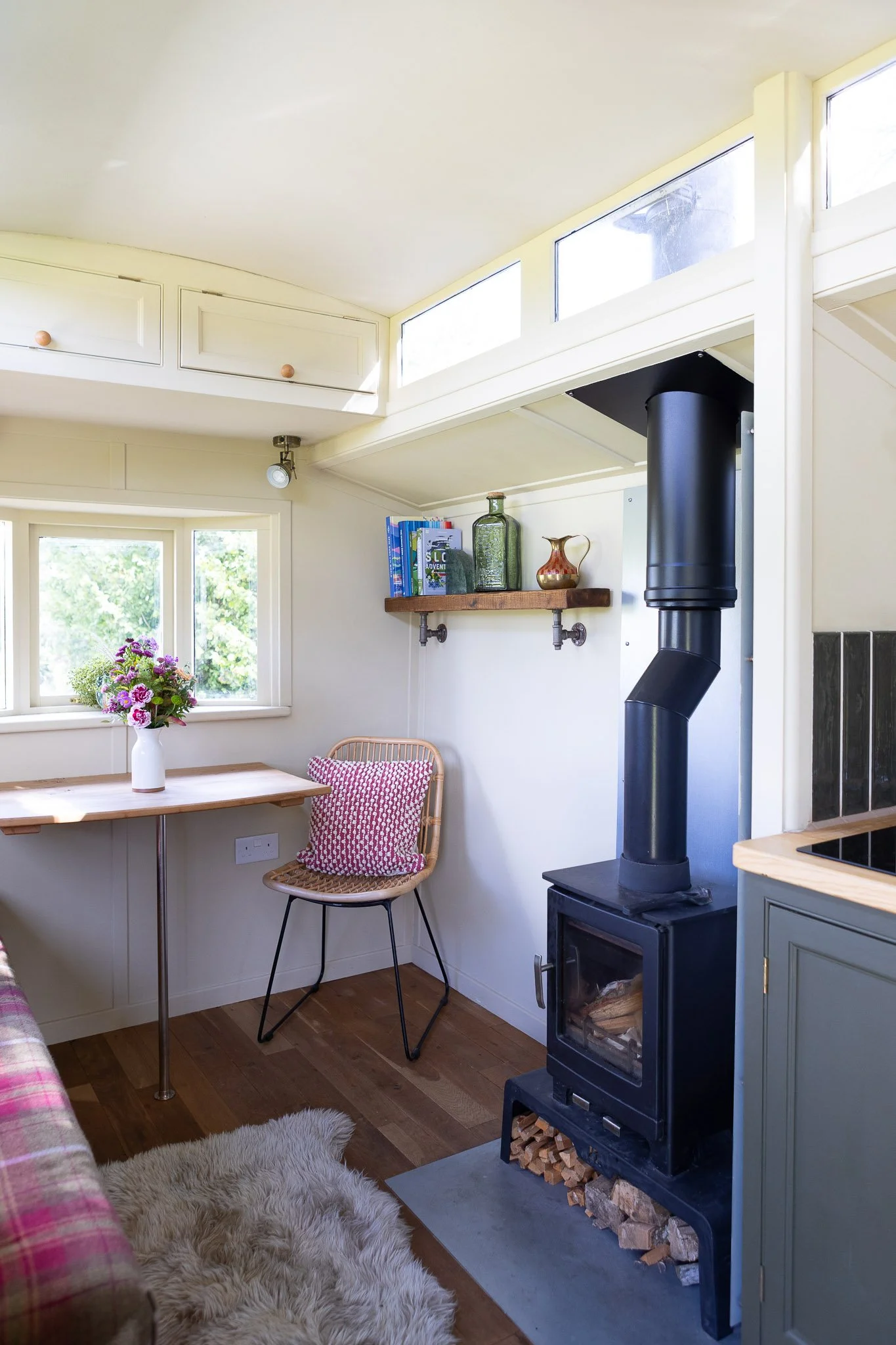 Cozy kitchen corner with a wood-burning stove, a wooden shelf with books and decorative bottles, a window with a vase of flowers on the table below, and a wool rug on a wooden floor.