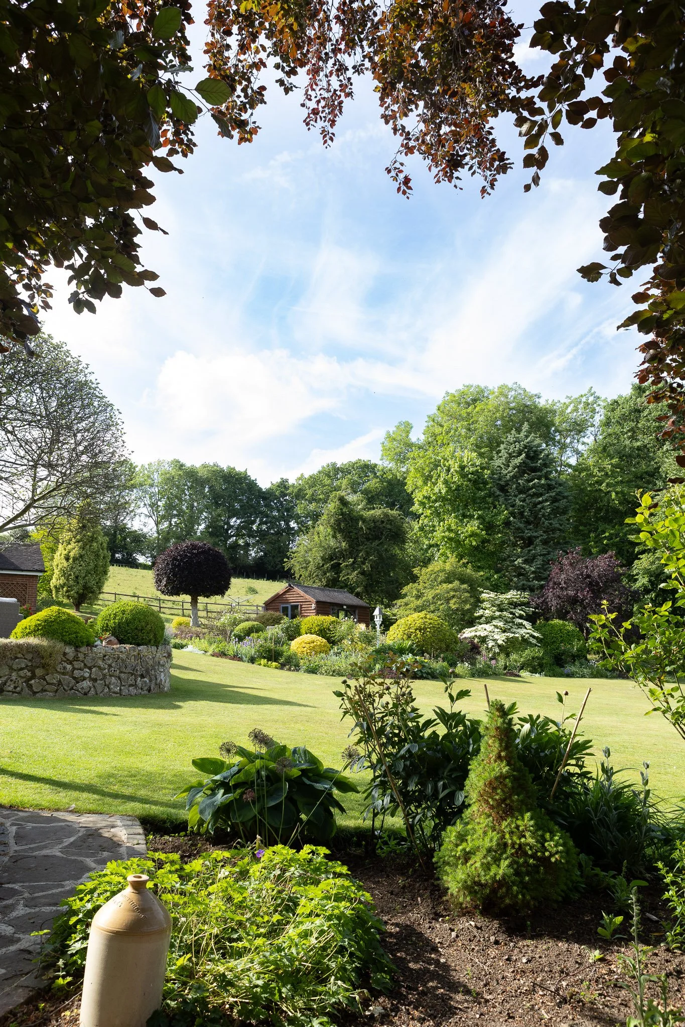 Lush garden with various green plants, bushes, and trees under a partly cloudy sky. Stone pathway and decorative pot in the foreground.