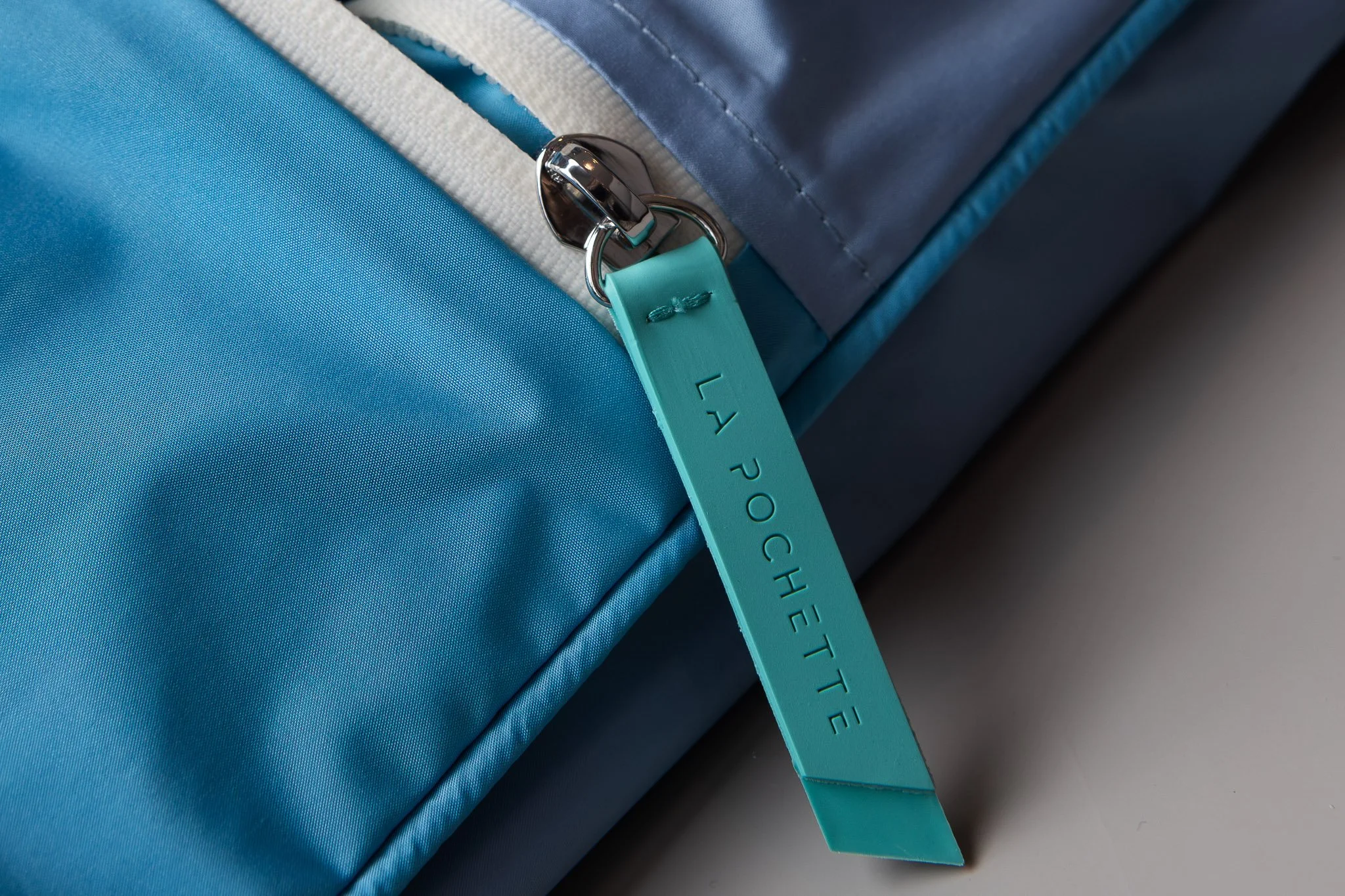 Close-up of a blue and teal fabric bag with a metal hook and a teal strap that has the word 'POCKET' embossed on it.