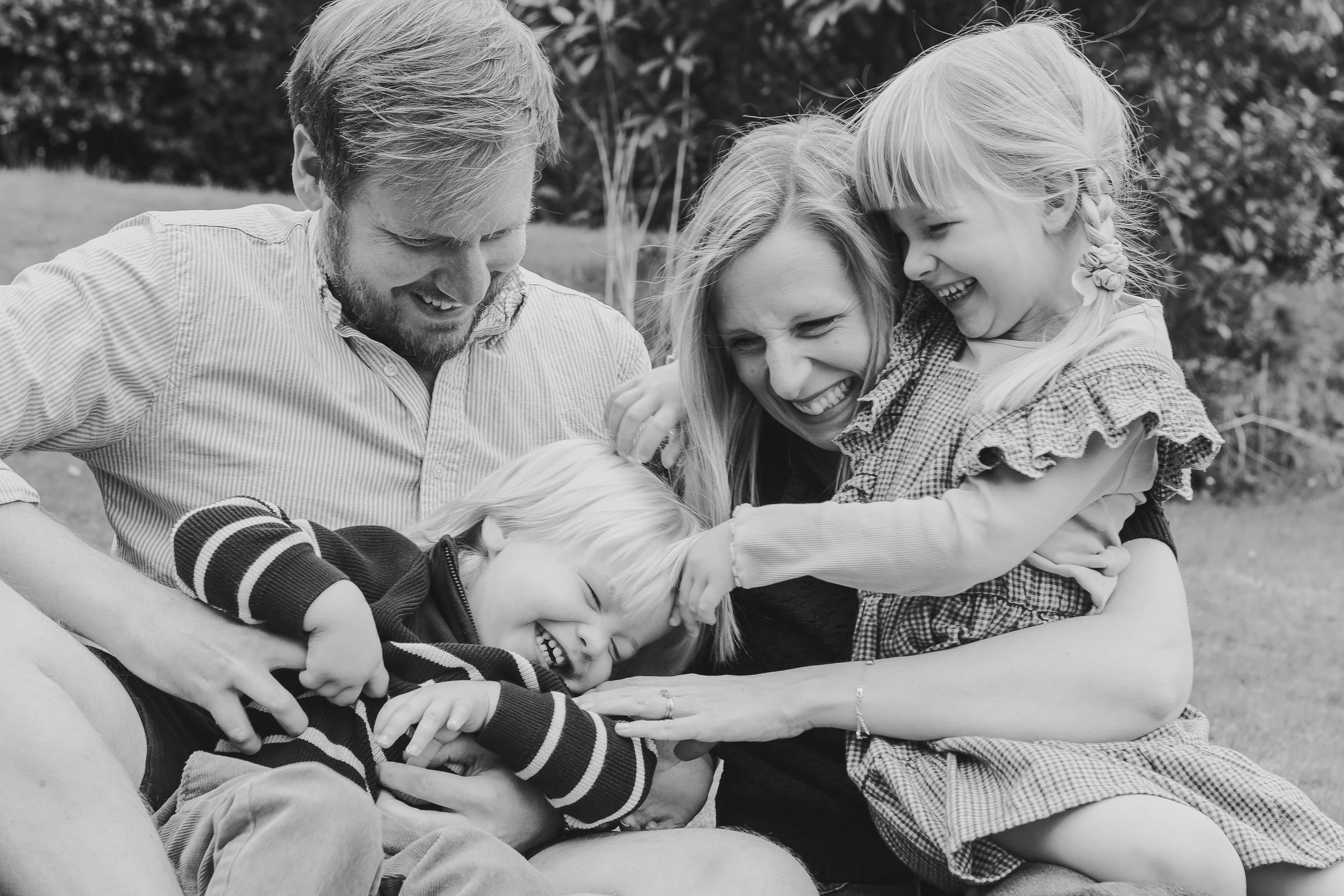 A family of five, including two children, a woman, and a man, sitting outdoors and laughing while holding and hugging each other in a joyful moment.