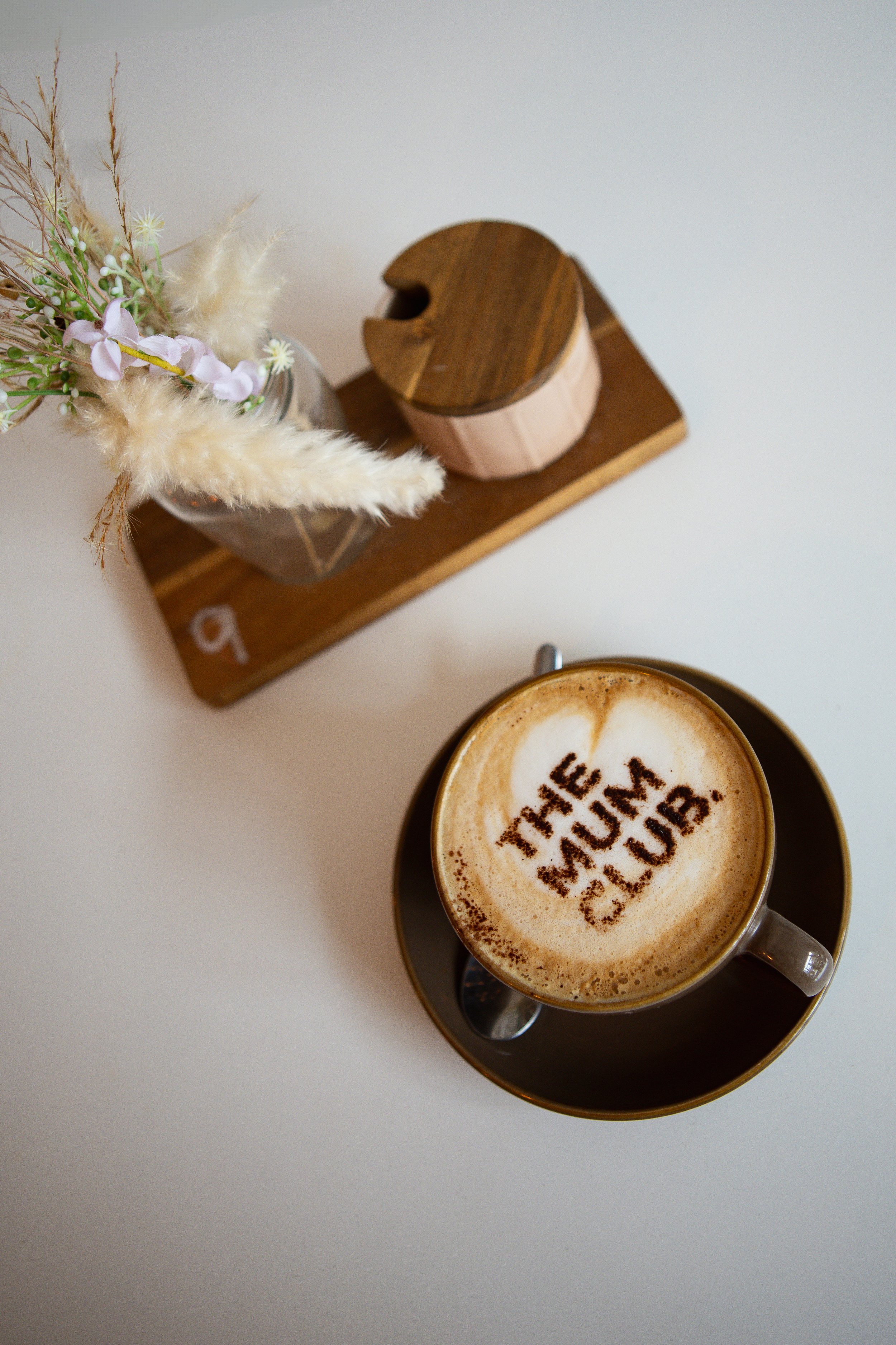 Top view of a coffee cup with foam and cinnamon powder inscription, placed on a saucer with a spoon, next to a wooden stand holding a small vase of dried flowers on a white surface.