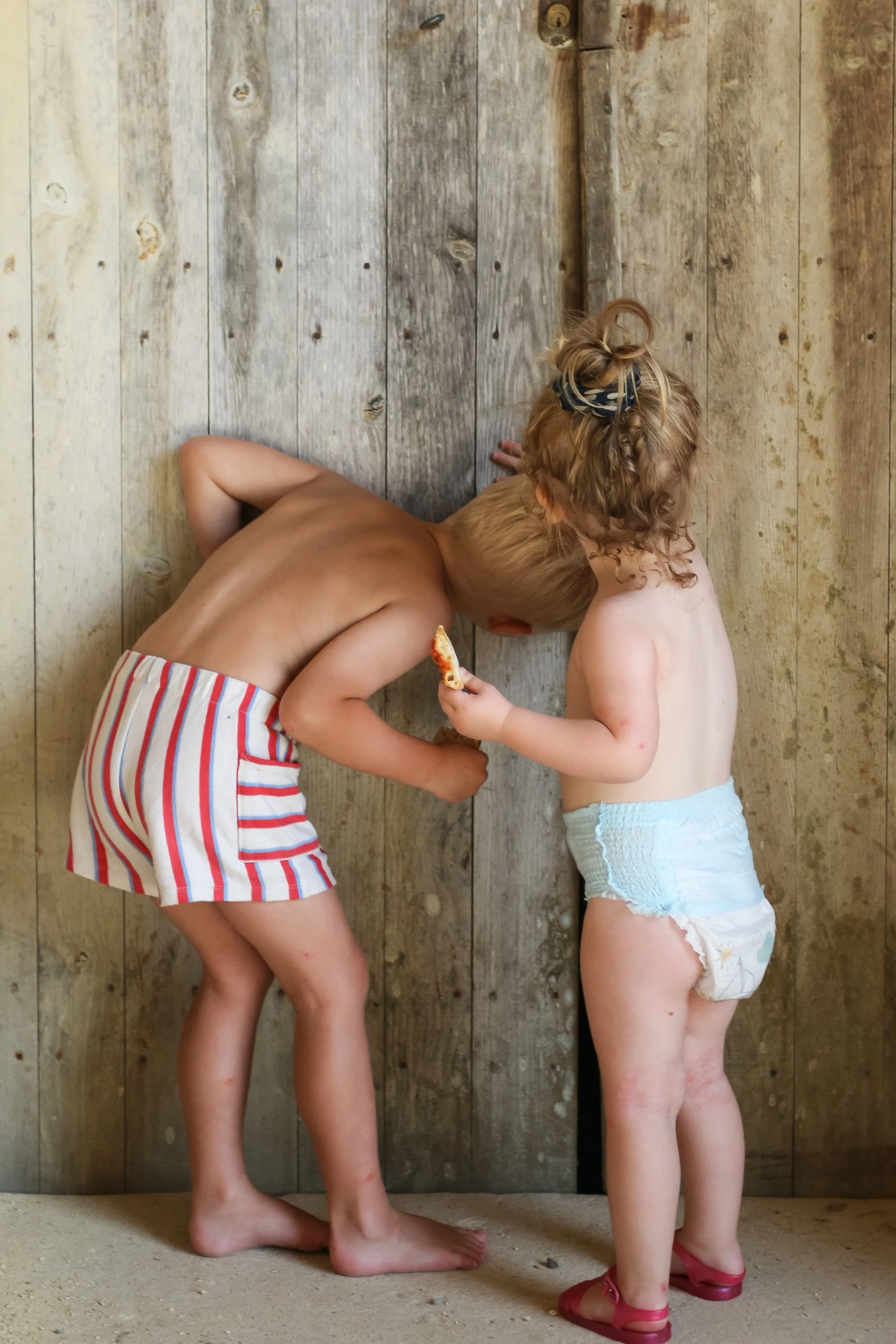 Two young children, a boy and a girl, are standing closely together facing a wooden wall. The boy has short blonde hair, is shirtless, and wearing striped shorts. The girl has curly hair tied with a bow, is only wearing a diaper and pink sandals, and