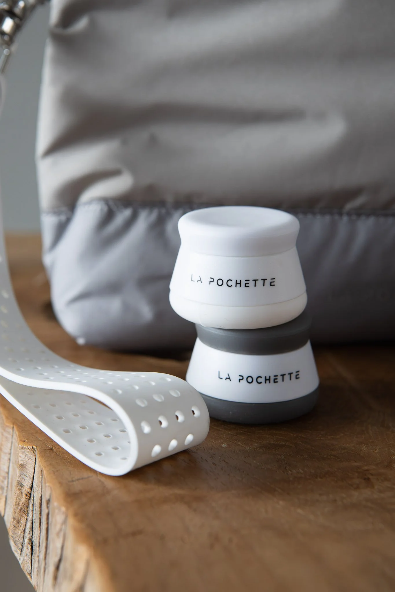 Close-up of two small white containers with black text, labeled 'LA POCHETTE,' placed on a wooden surface with a silver aluminum foil pouch and a white perforated silicone spoon nearby.