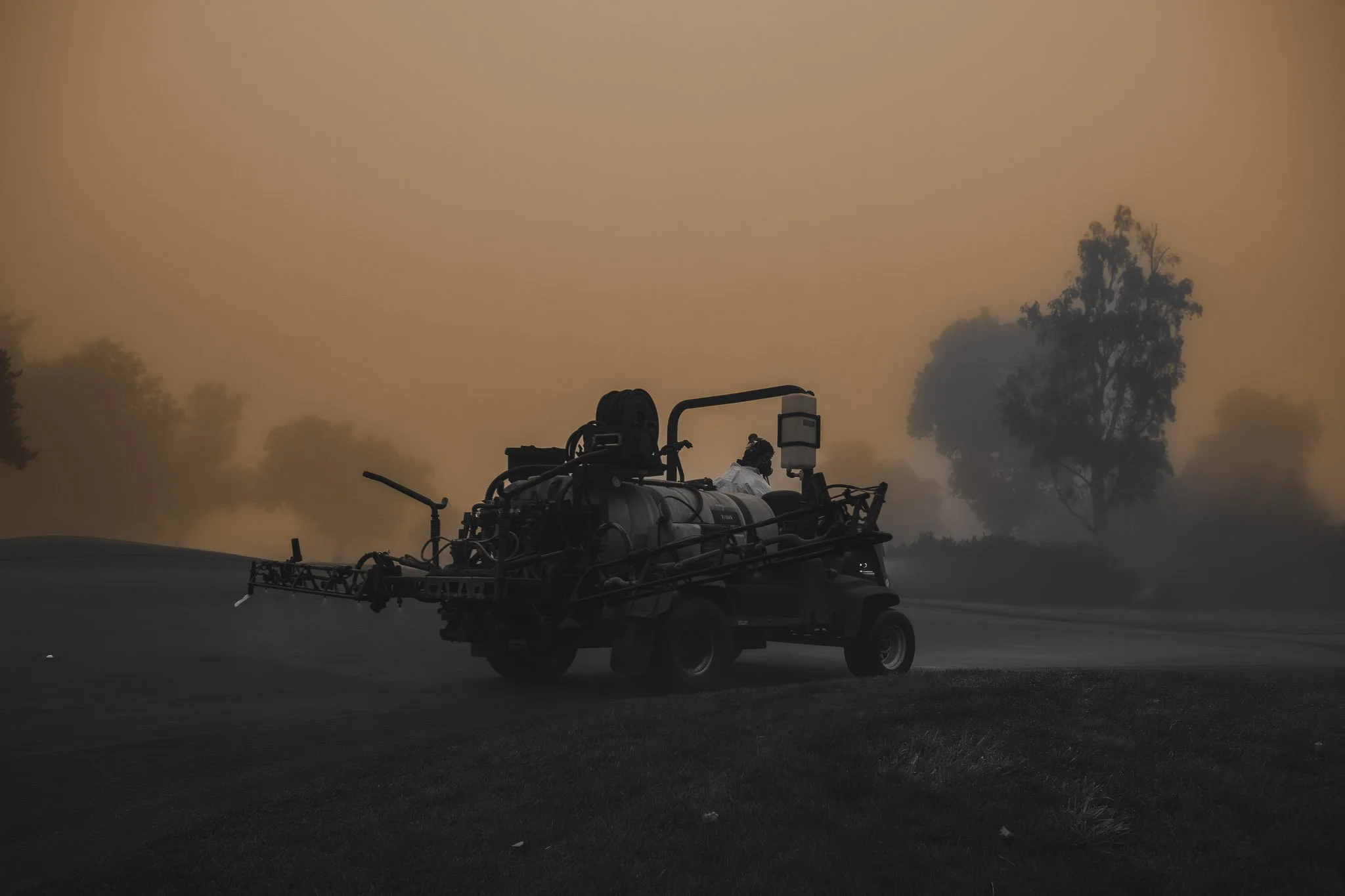 A street sweeper vehicle parked on a road with a smoky or foggy atmosphere, with silhouettes of trees in the background.