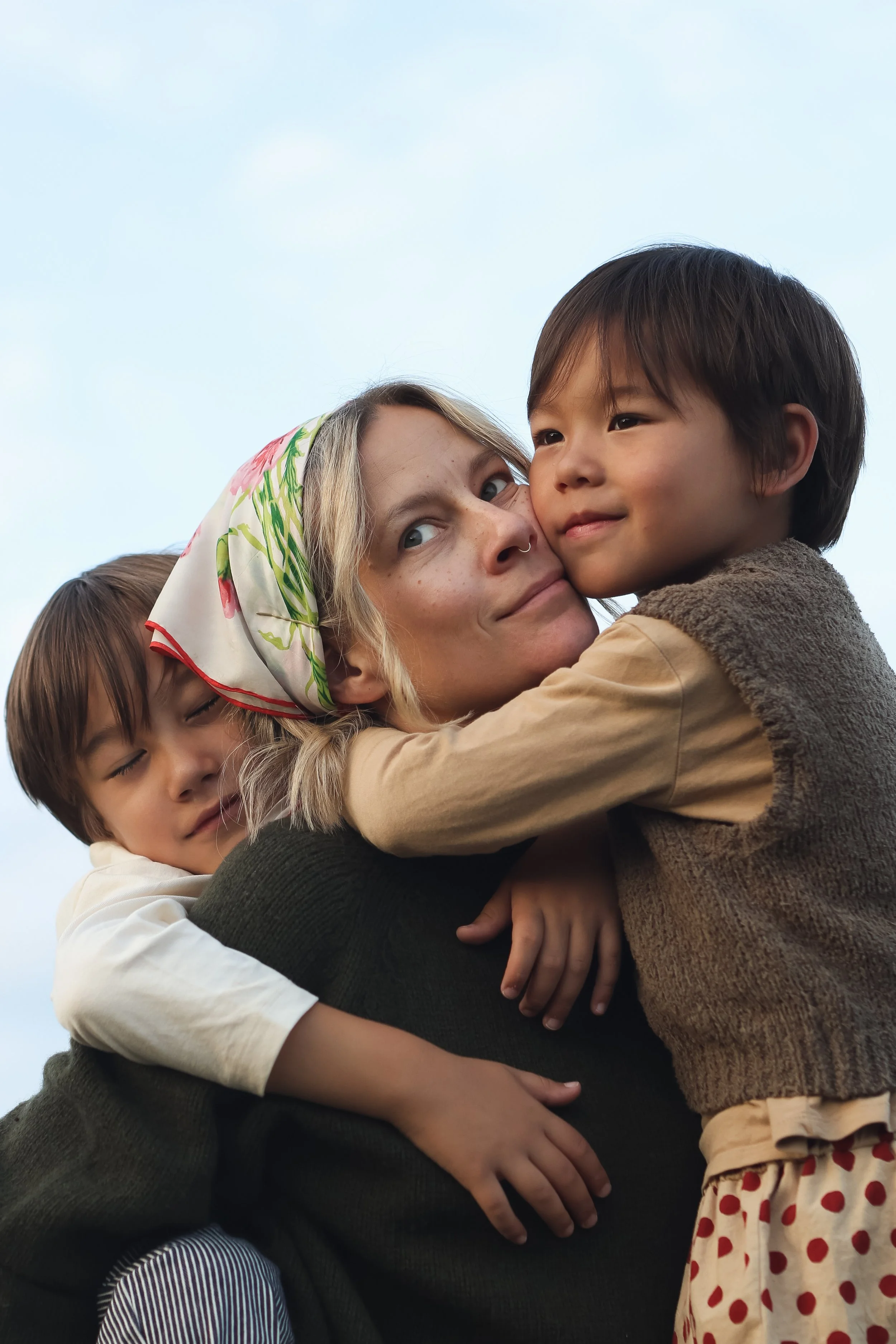 A woman with a floral headscarf hugging three children, one boy with dark hair, one girl with blond hair, and another child with dark hair and a striped shirt, against a clear sky.