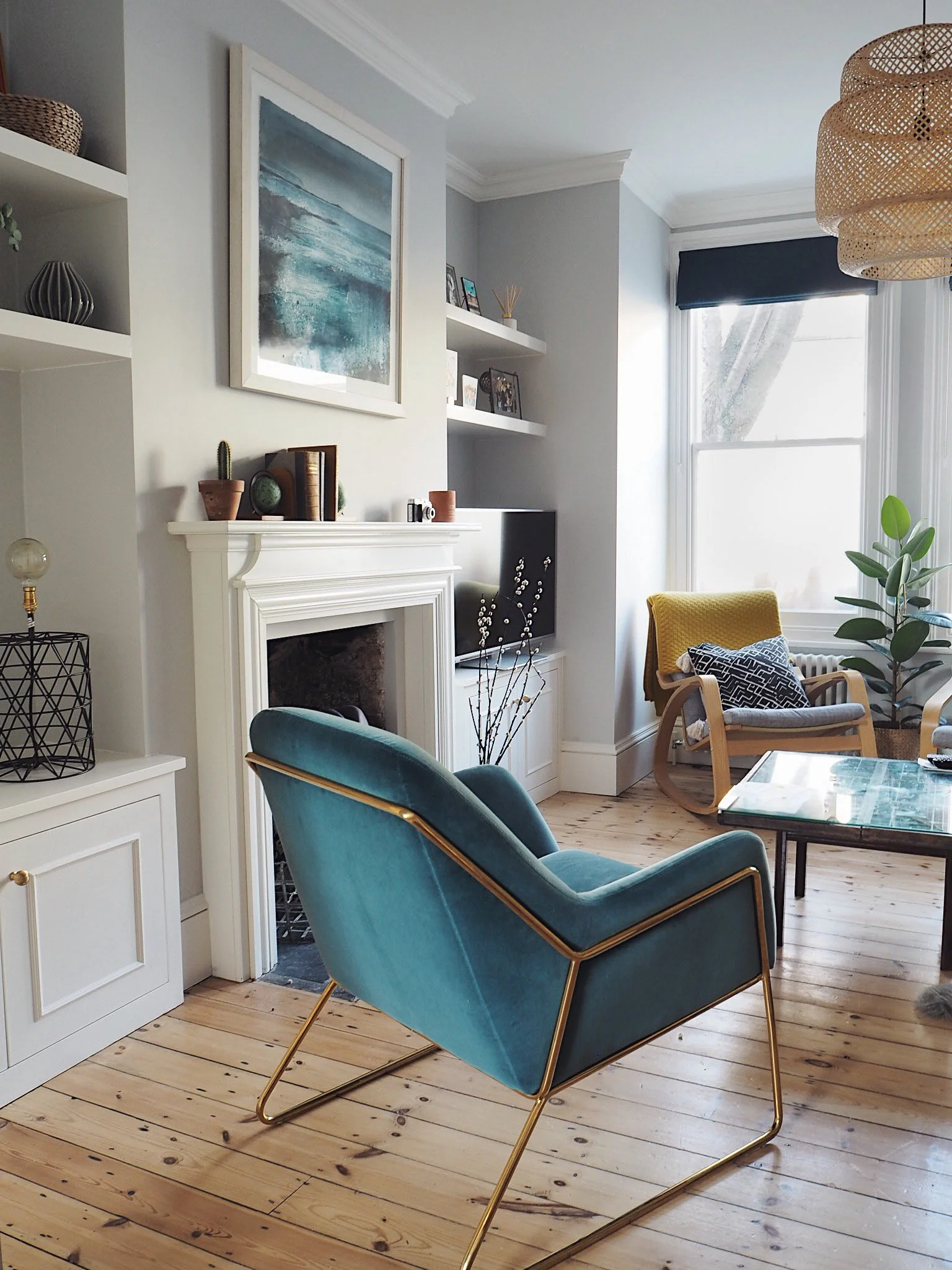 A cozy living room with a teal accent chair with gold legs, a fireplace with decorative items and artwork above, wooden flooring, and a large window with light curtains letting in natural light.