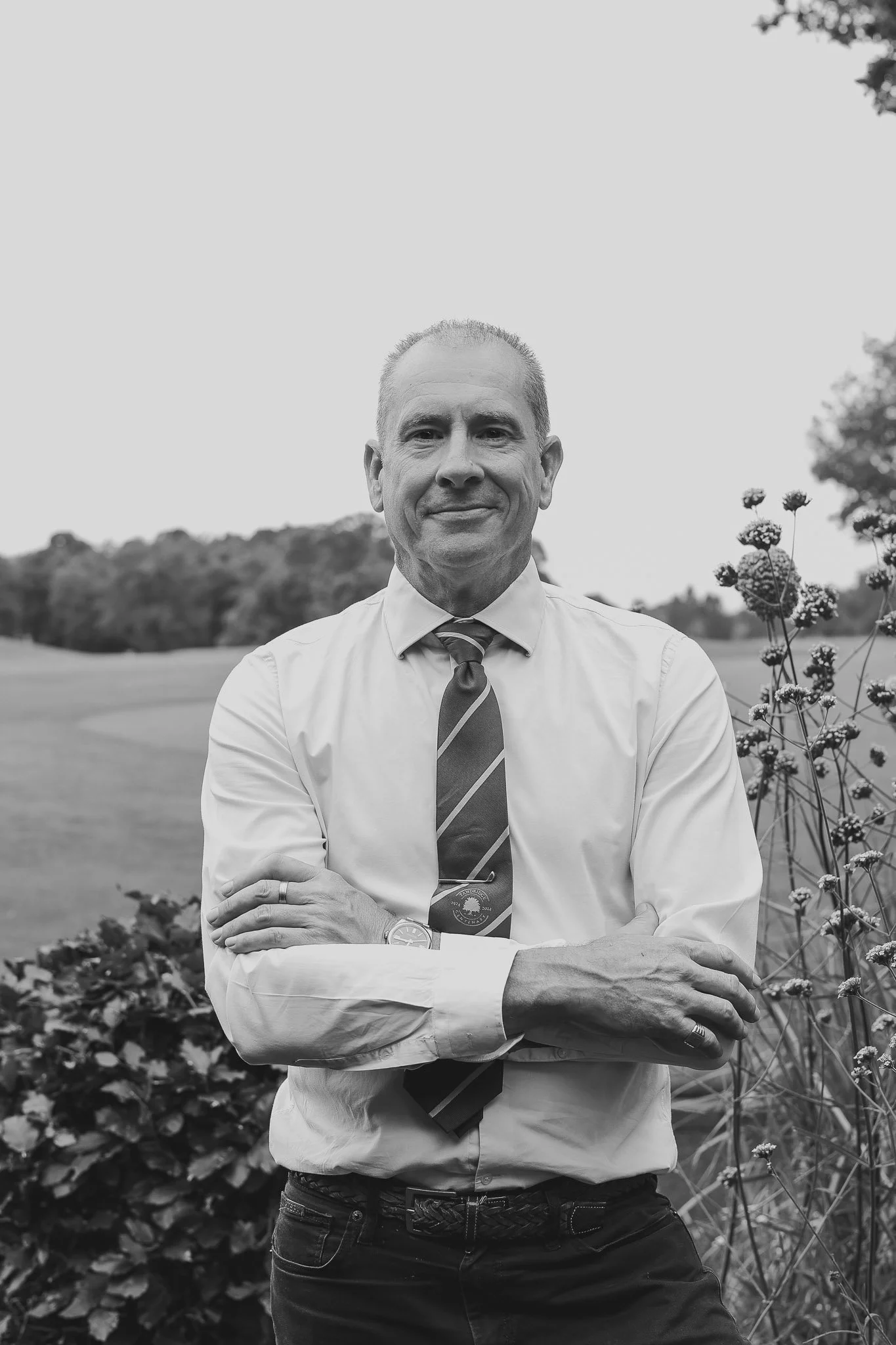 Black and white photo of a man standing outdoors in front of trees and flowers, wearing a dress shirt, tie, and a watch, with arms crossed and a slight smile.