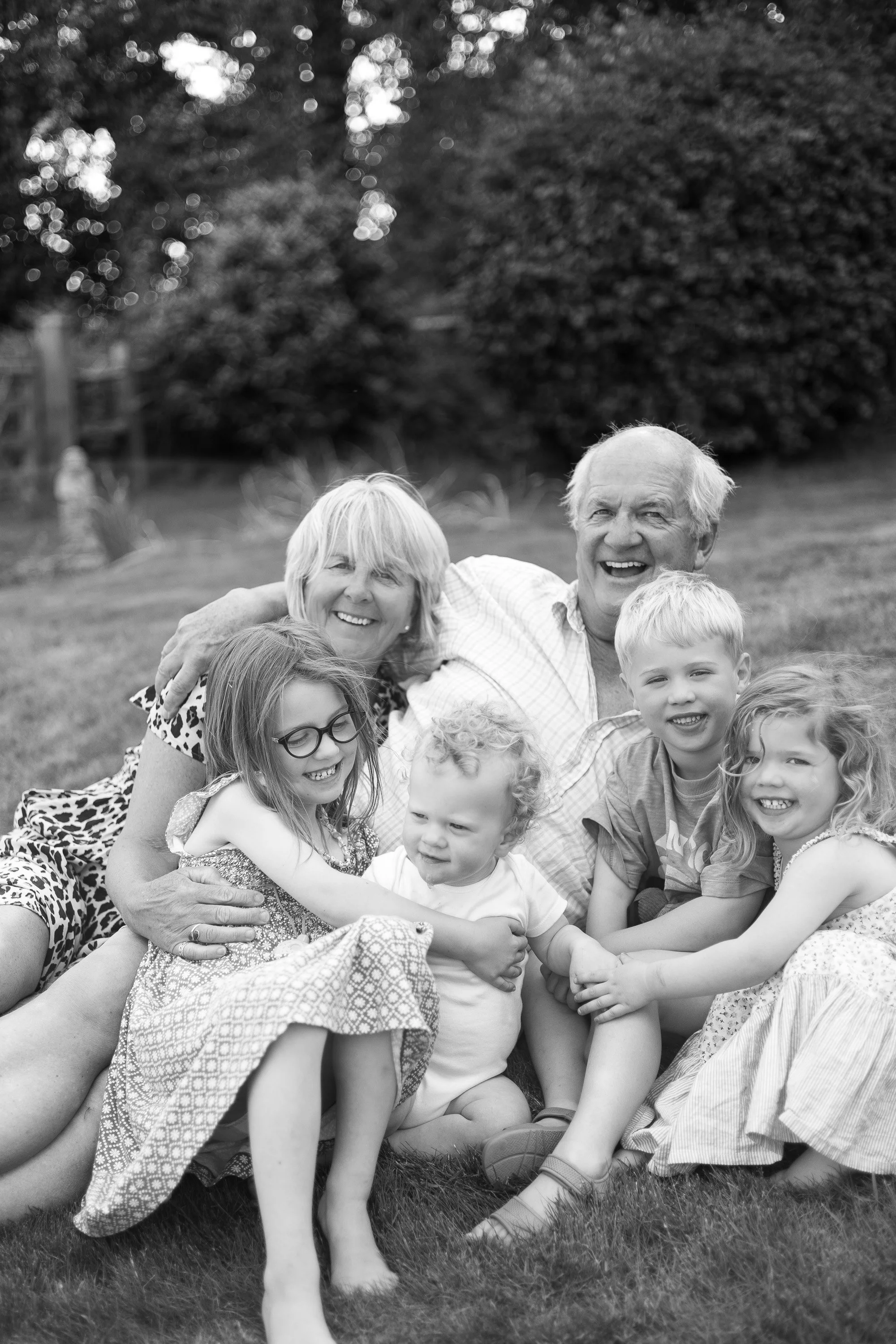 A black and white photo of a family of seven, including children and grandparents, smiling and hugging outdoors on a grassy field with trees in the background.