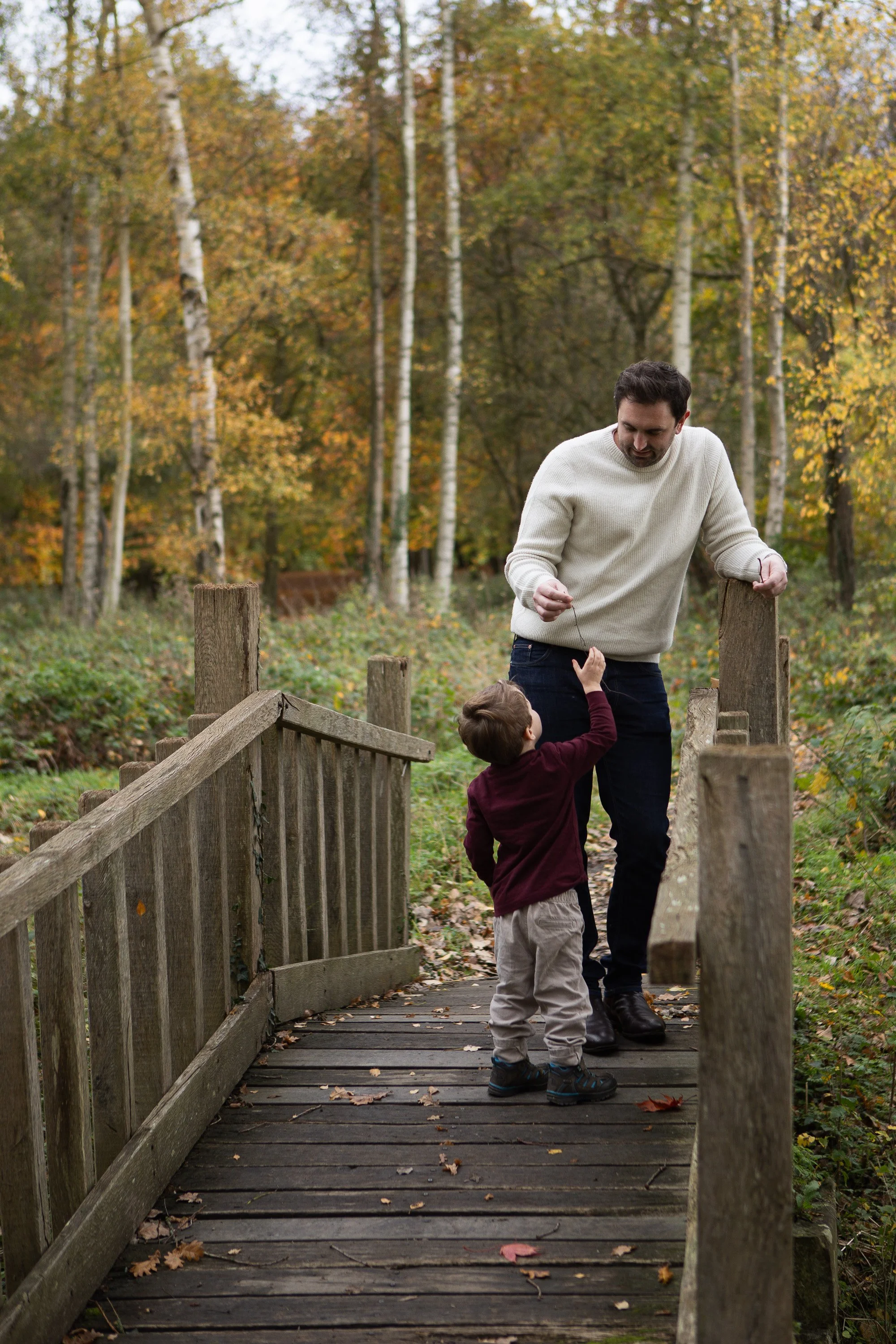 A man and a young boy walk together on a wooden footbridge surrounded by autumn trees in a forest.