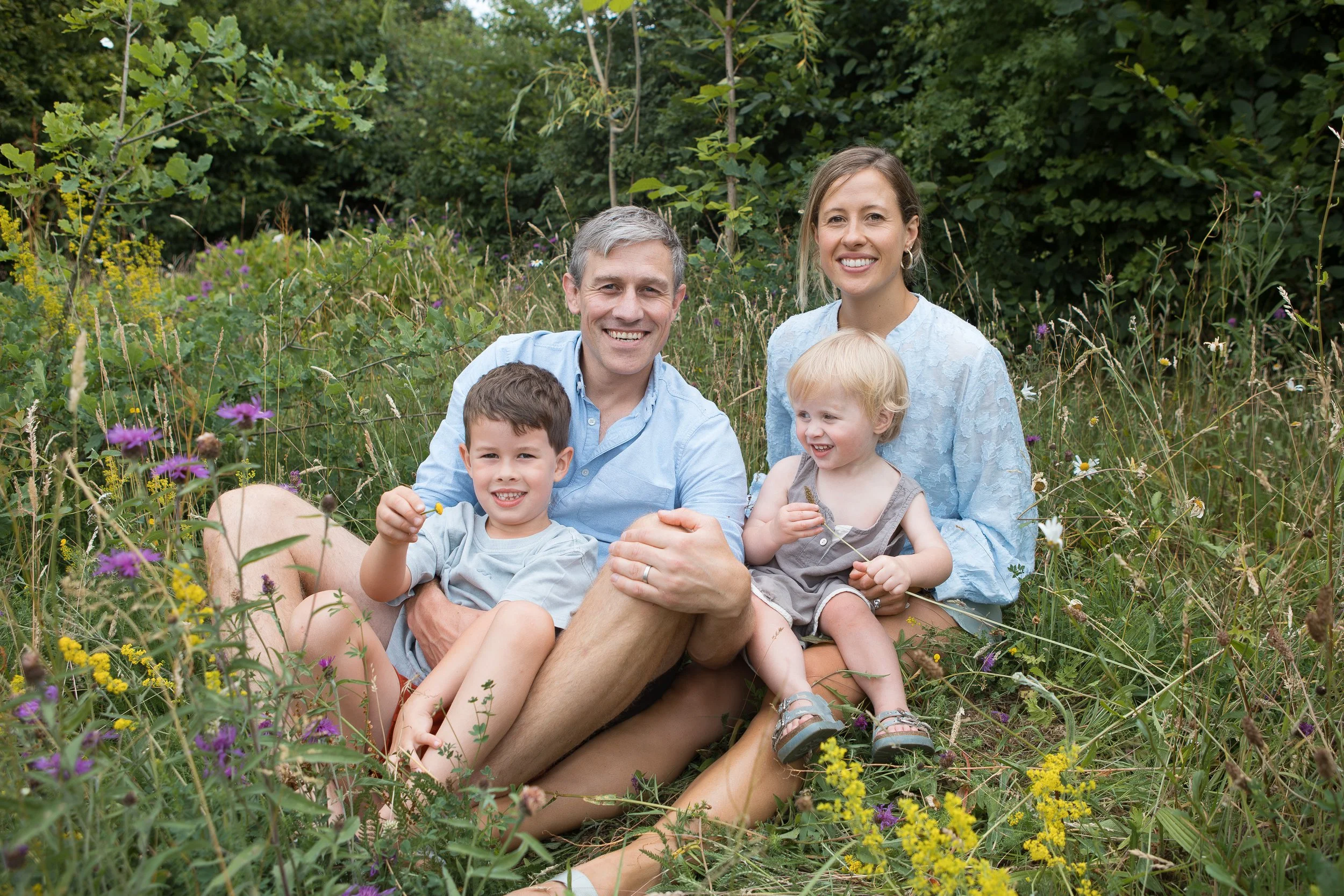 A happy family of four sitting in a field of wildflowers and greenery, outdoors during daytime. The father, mother, and two children are smiling, with the father holding the young boy and the mother holding the young girl.