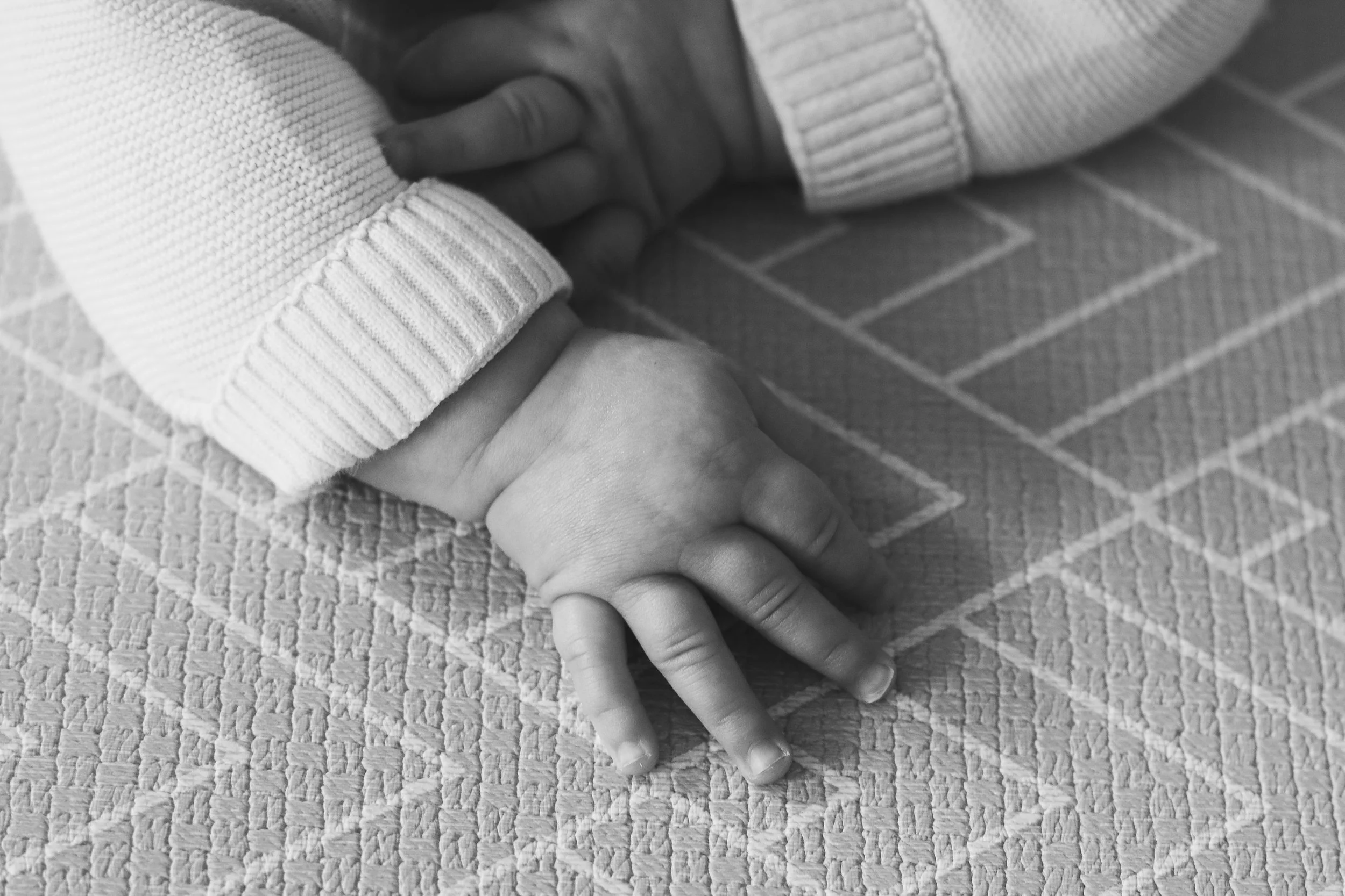 Close-up of a person's hand holding another person's hand, with the person receiving support wearing a light-colored, ribbed sweater, on a textured, patterned surface.