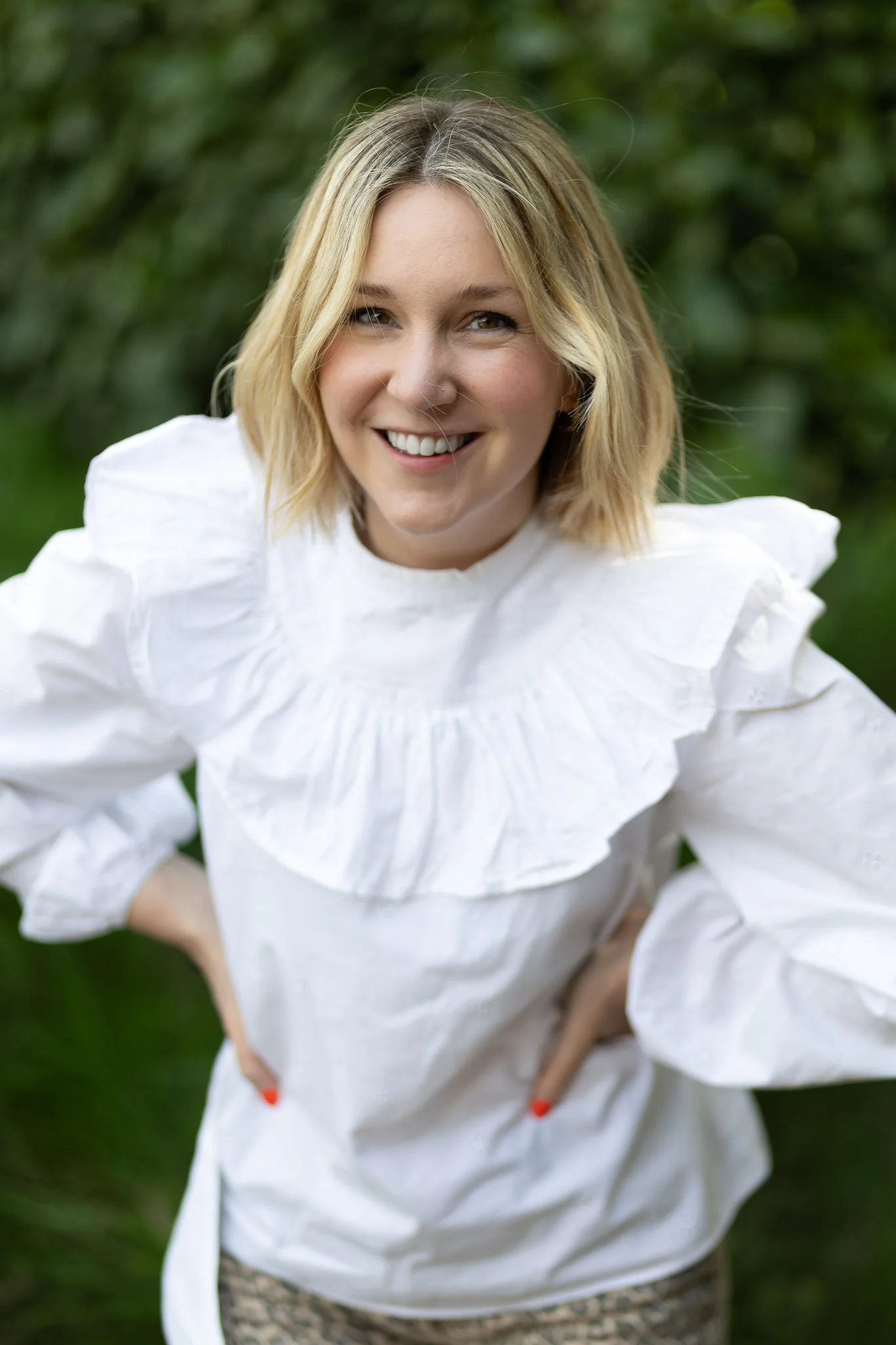 A smiling woman with blonde hair wearing a white top standing outdoors with greenery in the background.