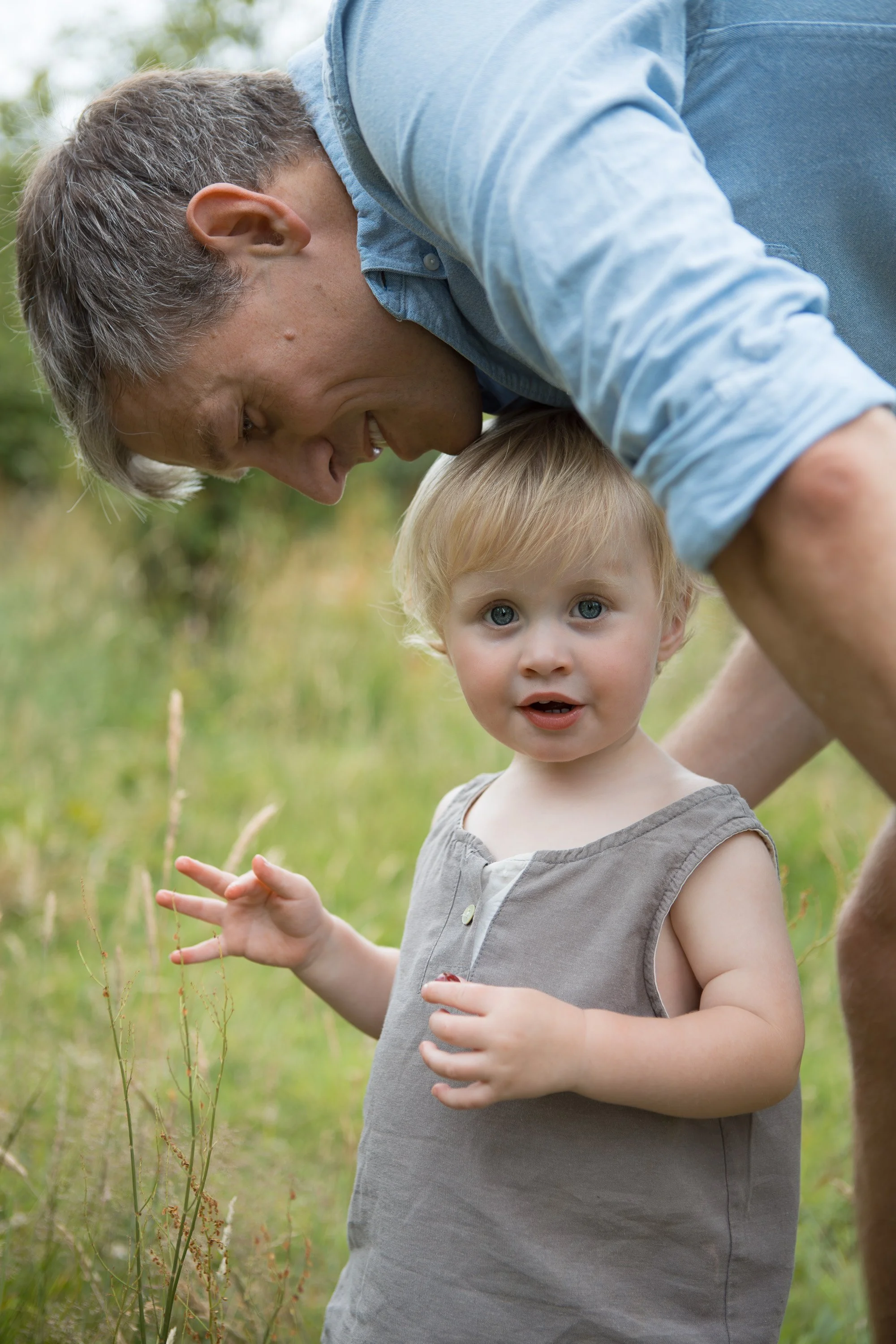 A man and a young girl outdoors in a grassy area, with the man leaning over and the girl looking at the camera, holding a dandelion.
